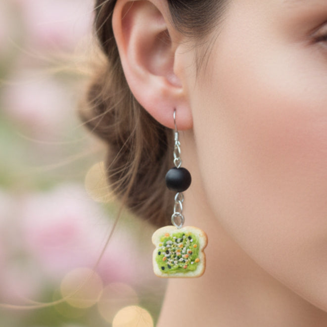 Pair of earrings shaped like slices of bread with green spread on a pink background