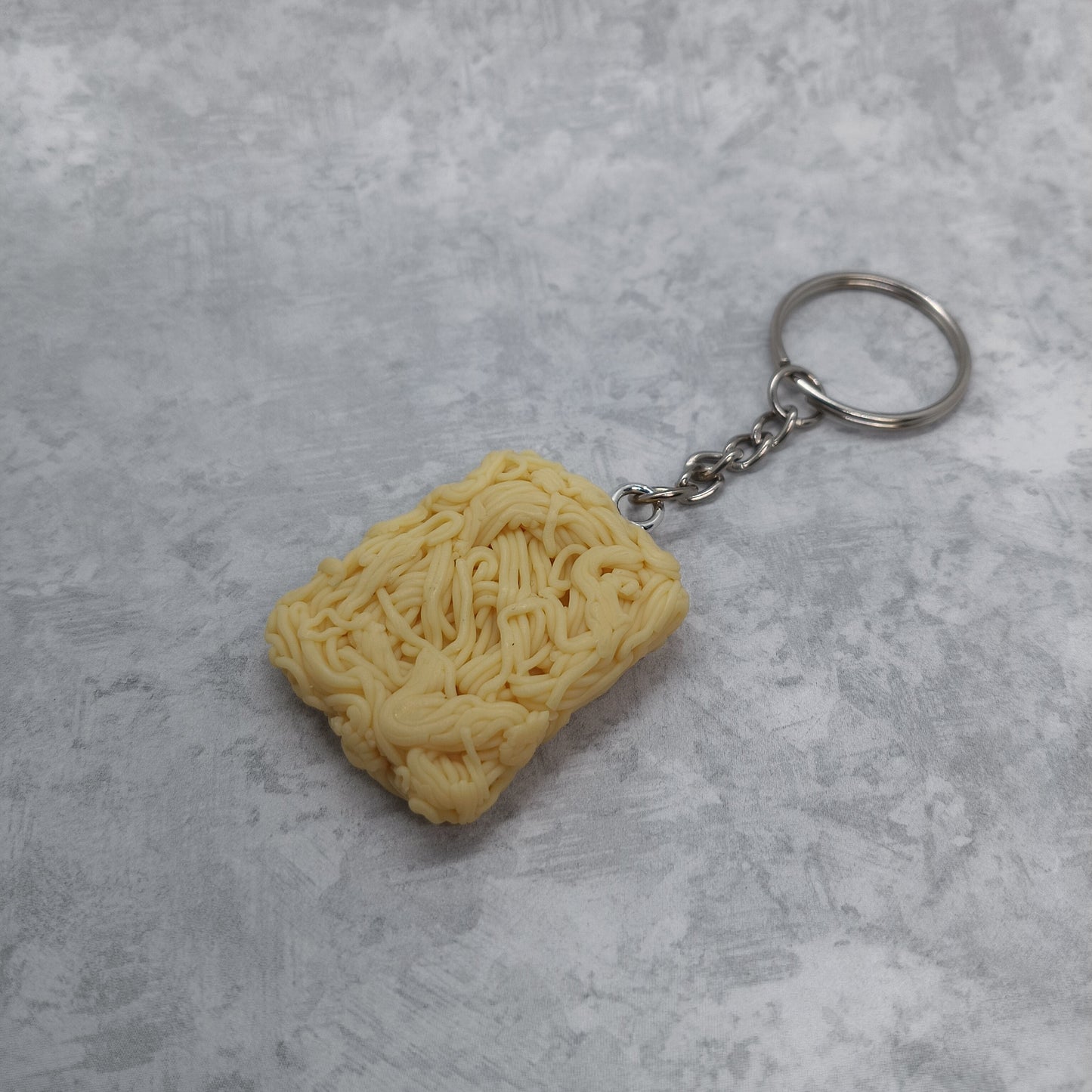 A novelty keychain shaped like a block of instant ramen noodles, attached to a silver metal chain and split ring, displayed on a textured gray background.