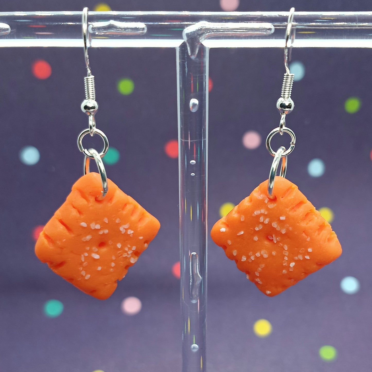 A pair of orange square dangle earrings with white salt-like speckles, hanging from silver fishhook wires on a clear acrylic display stand against a dark purple background with colorful confetti.