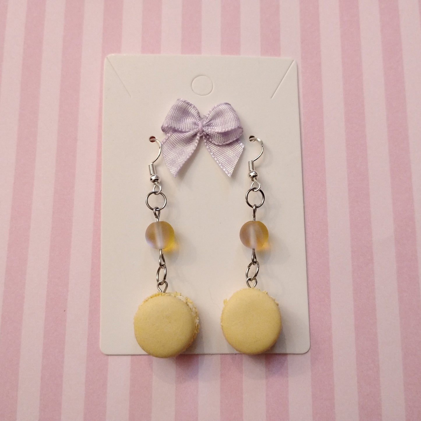 Yellow macaron earrings with a purple bow on a striped pink background