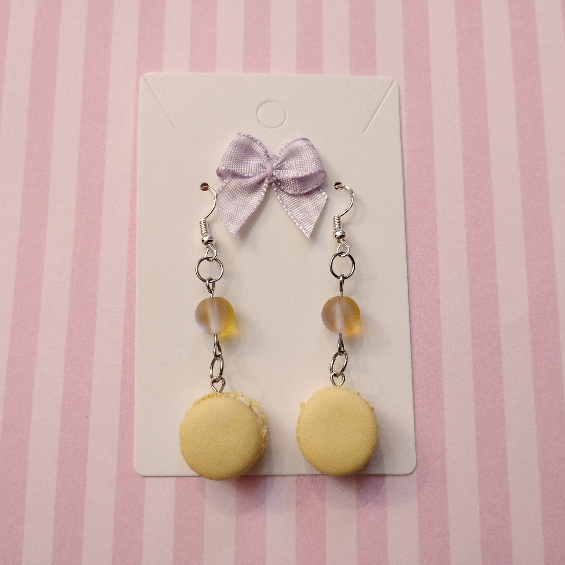 Yellow macaron earrings with a purple bow on a striped pink background