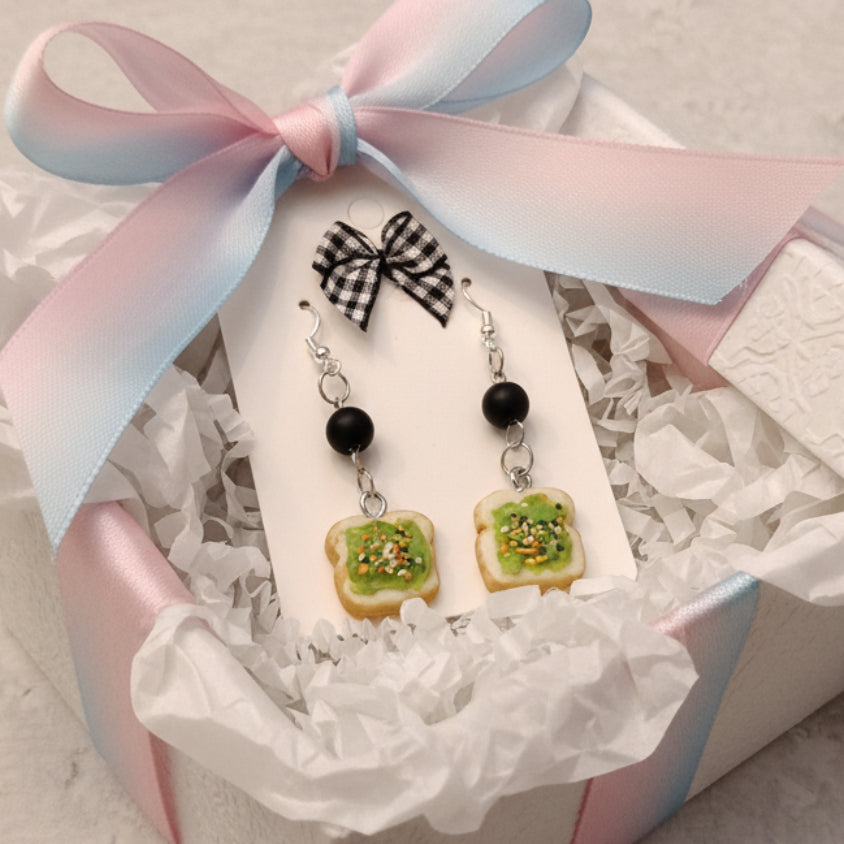 Pair of earrings resembling toast with avocado on a card against a gray background