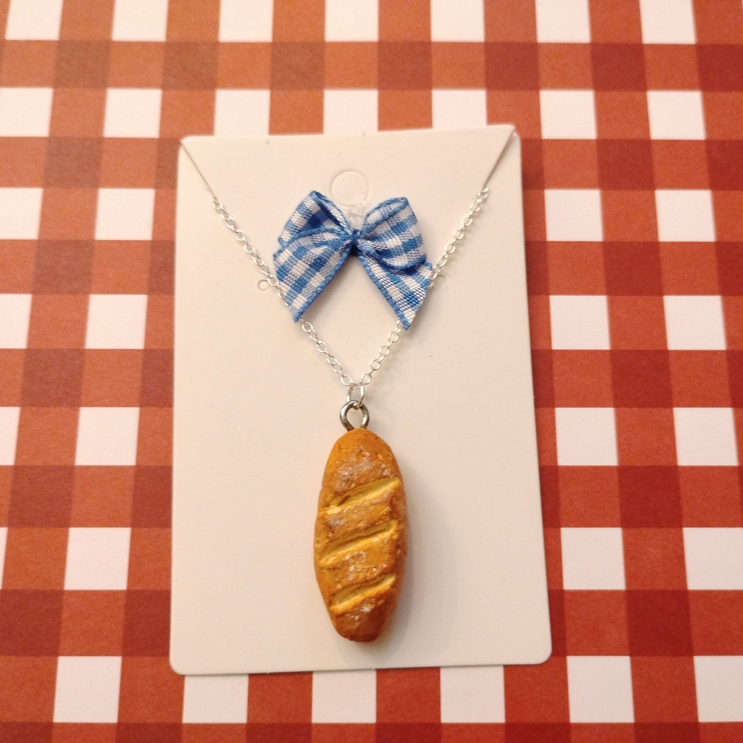 Necklace with bread-shaped pendant and blue checkered bow on a red and white checkered background