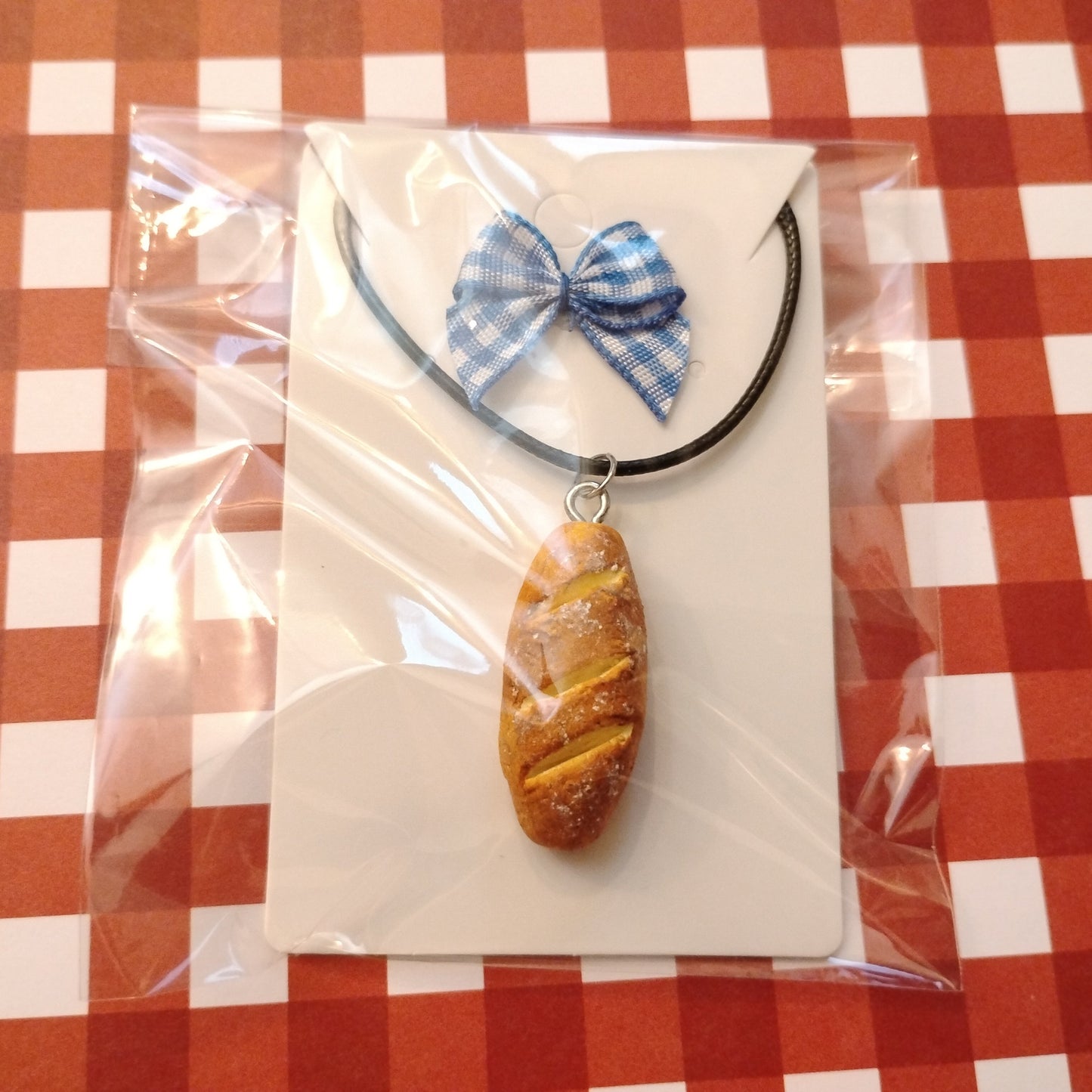 Pendant necklace with a blue bow and bread-shaped charm in clear packaging on a red and white checkered tablecloth.