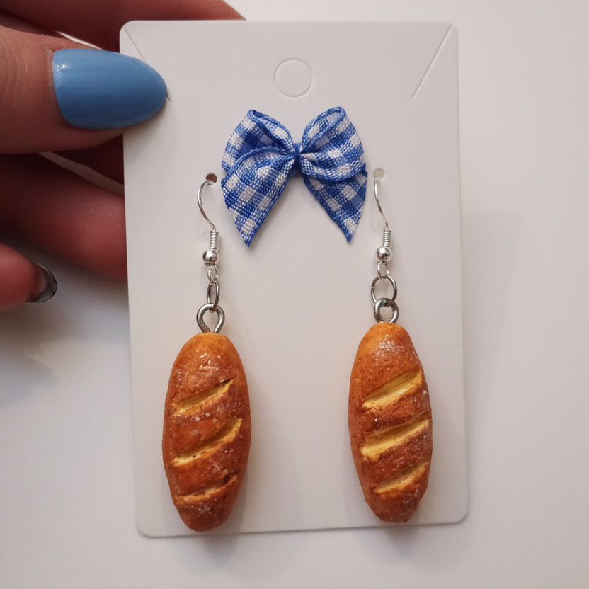 Bread-shaped earrings with a blue bow on a white card, held by a hand.
