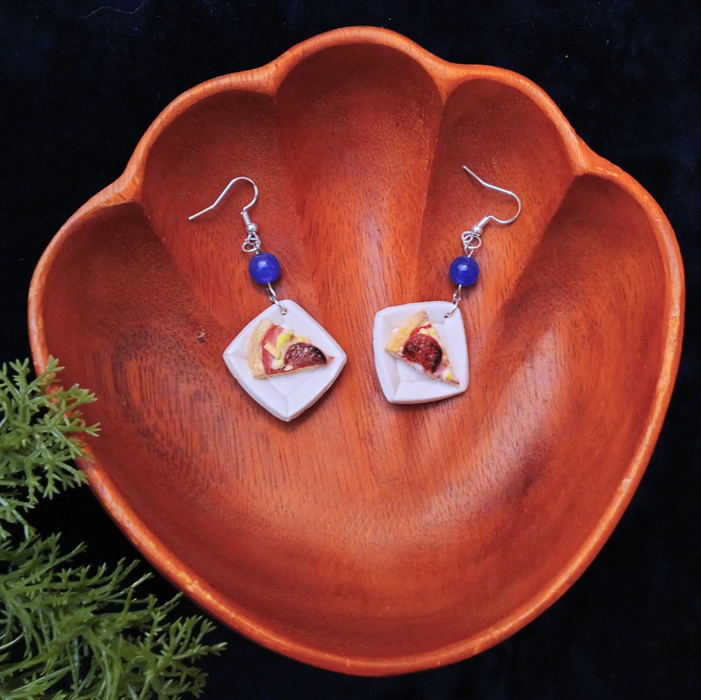 Pair of earrings shaped like pizza slices with blue beads on a wooden dish.