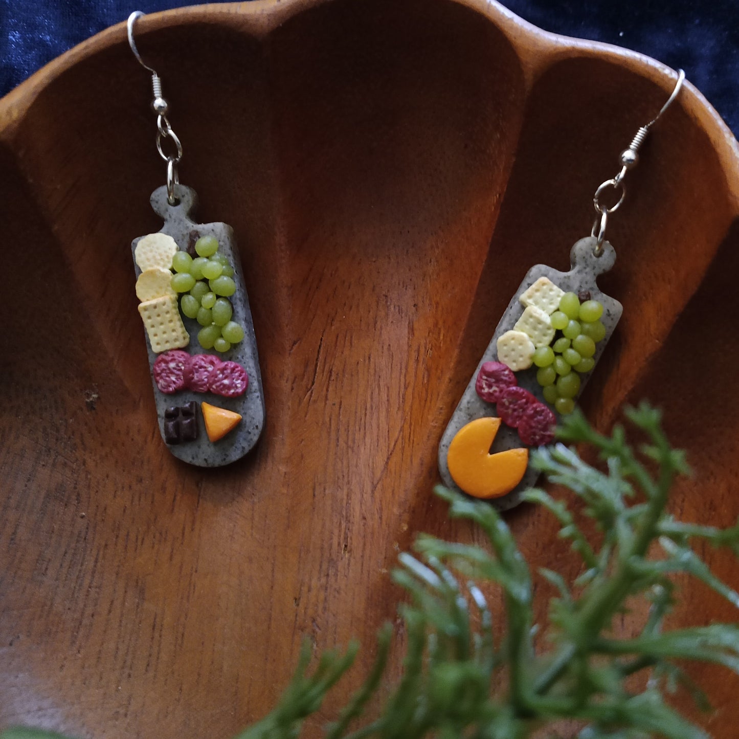 Pair of earrings resembling a charcuterie board on a wooden surface with a plant in the foreground.