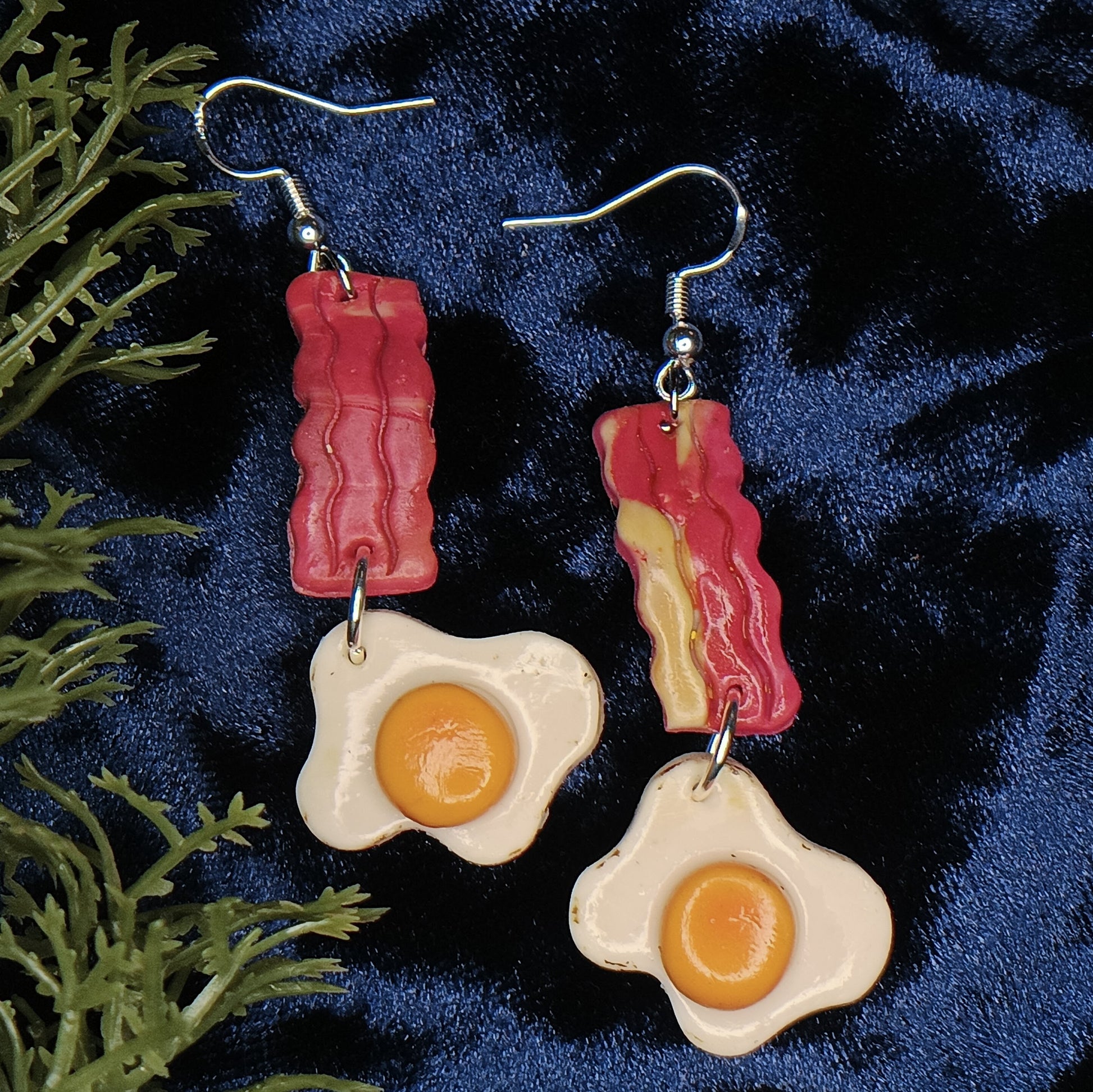 Bacon and egg earrings on a dark blue fabric background with green leaves.