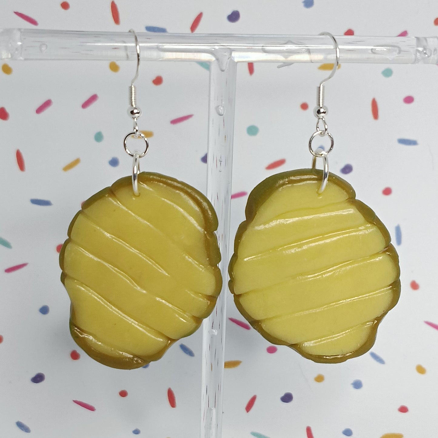 A pair of olive-yellow resin earrings shaped like stylized pickle slices, hanging from silver fishhook wires on a clear display stand against a white background with colorful confetti dots.