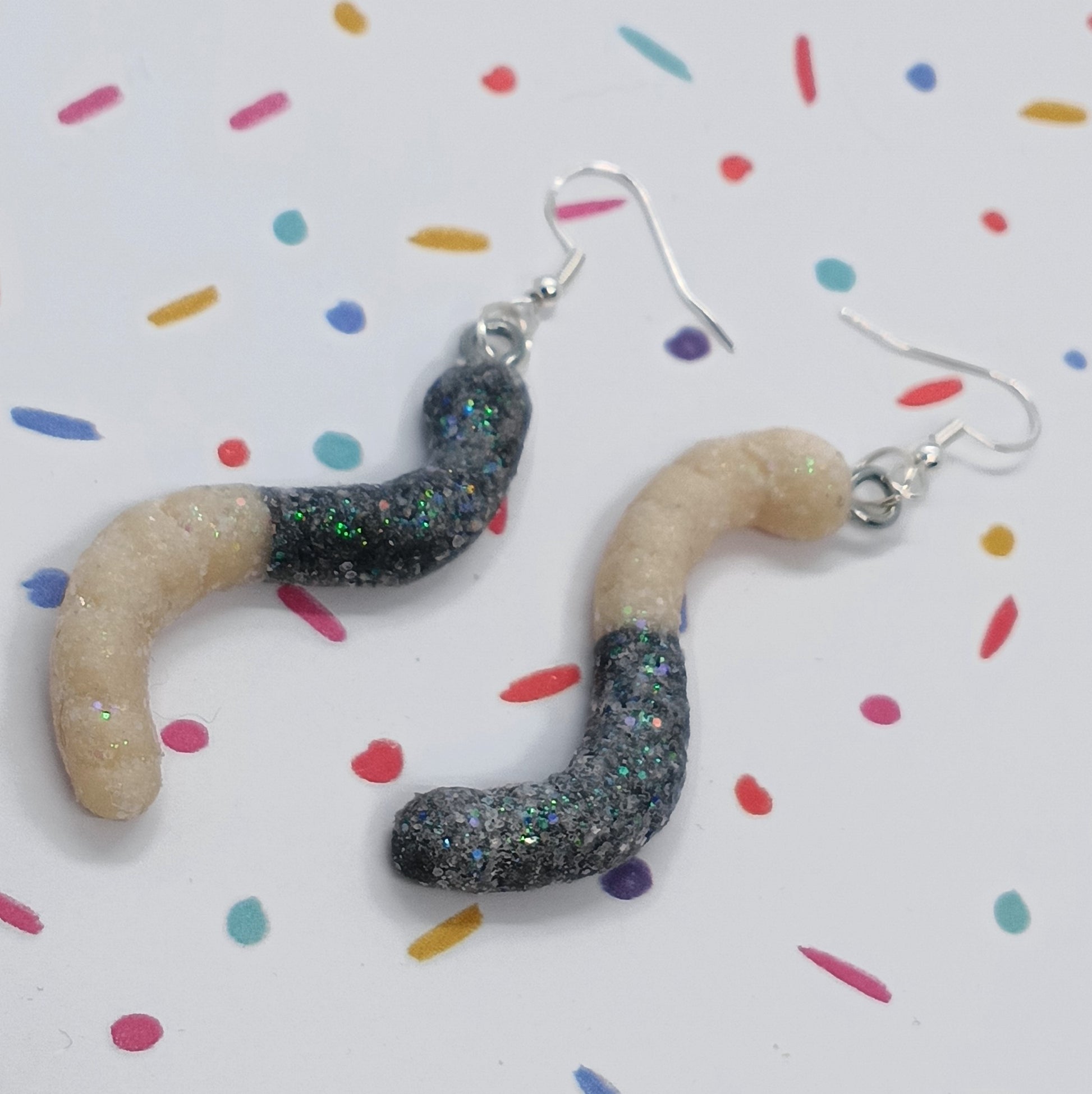 A pair of handmade worm-shaped dangle earrings with a beige and glittery black design, hanging from silver fishhook wires on a white background with colorful confetti.