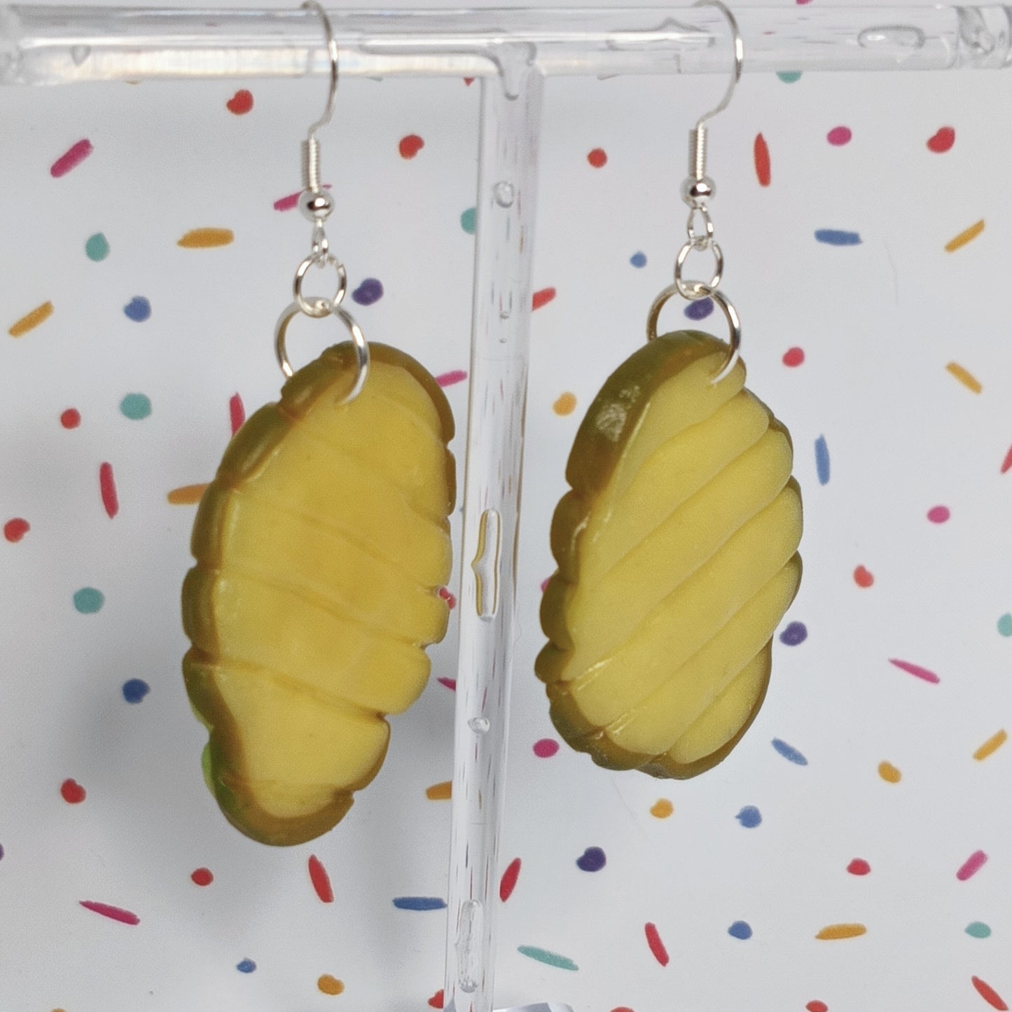 A pair of novelty earrings shaped like pickle slices, hanging from silver fishhook wires on a clear acrylic display stand against a white background with colorful confetti dots.