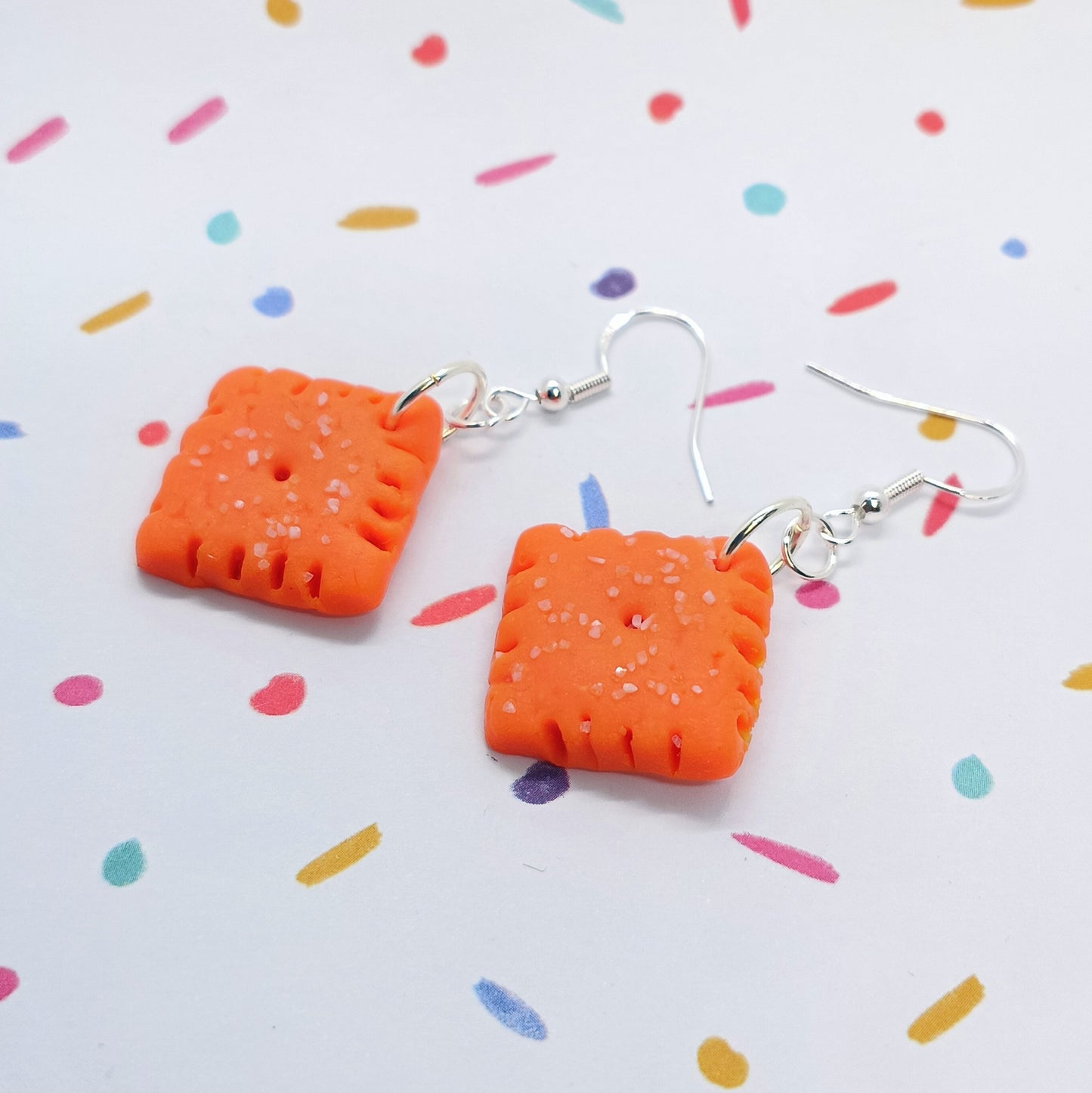A pair of orange square-shaped dangle earrings with textured details and silver fishhook wires, displayed on a white background with colorful confetti.