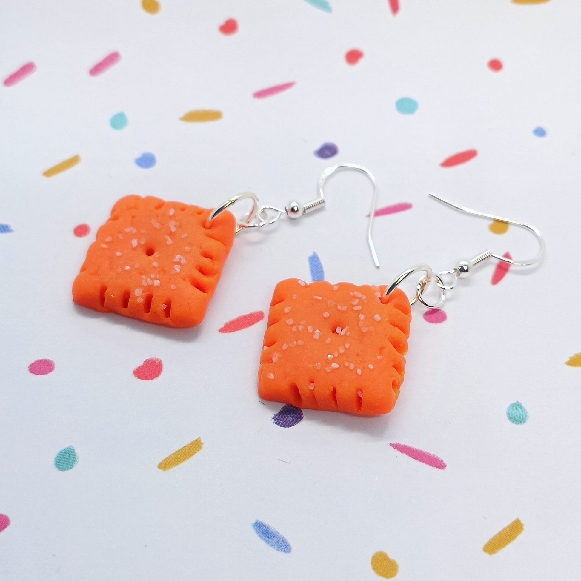 A pair of orange square-shaped dangle earrings with textured details and silver fishhook wires, displayed on a white background with colorful confetti.