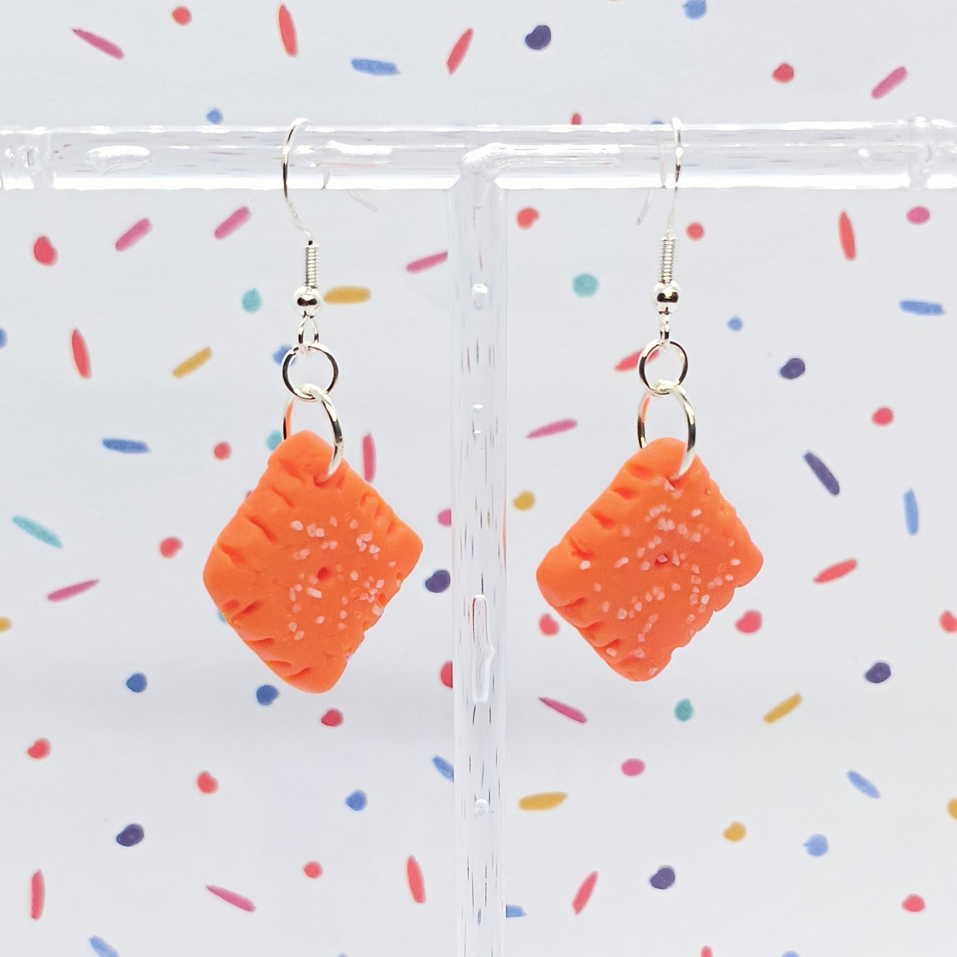 A pair of orange square dangle earrings with embossed texture and small white speckles, hanging from silver fishhook wires on a clear acrylic display stand against a white background with colorful confetti.