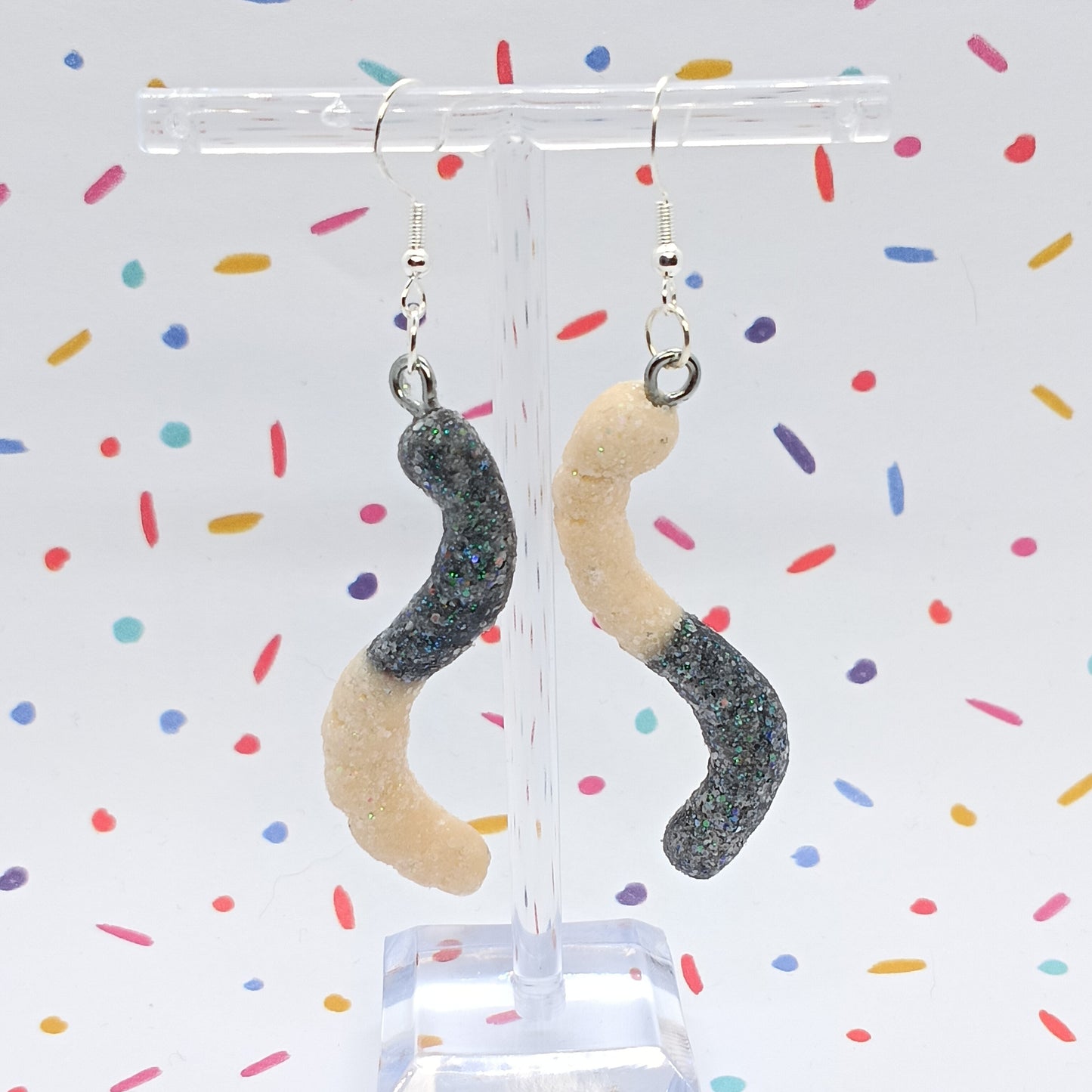 A pair of dangling earrings shaped like sour gummy worms, with glittery dark gray and beige sections, hanging from a clear acrylic display stand against a white background with colorful confetti.