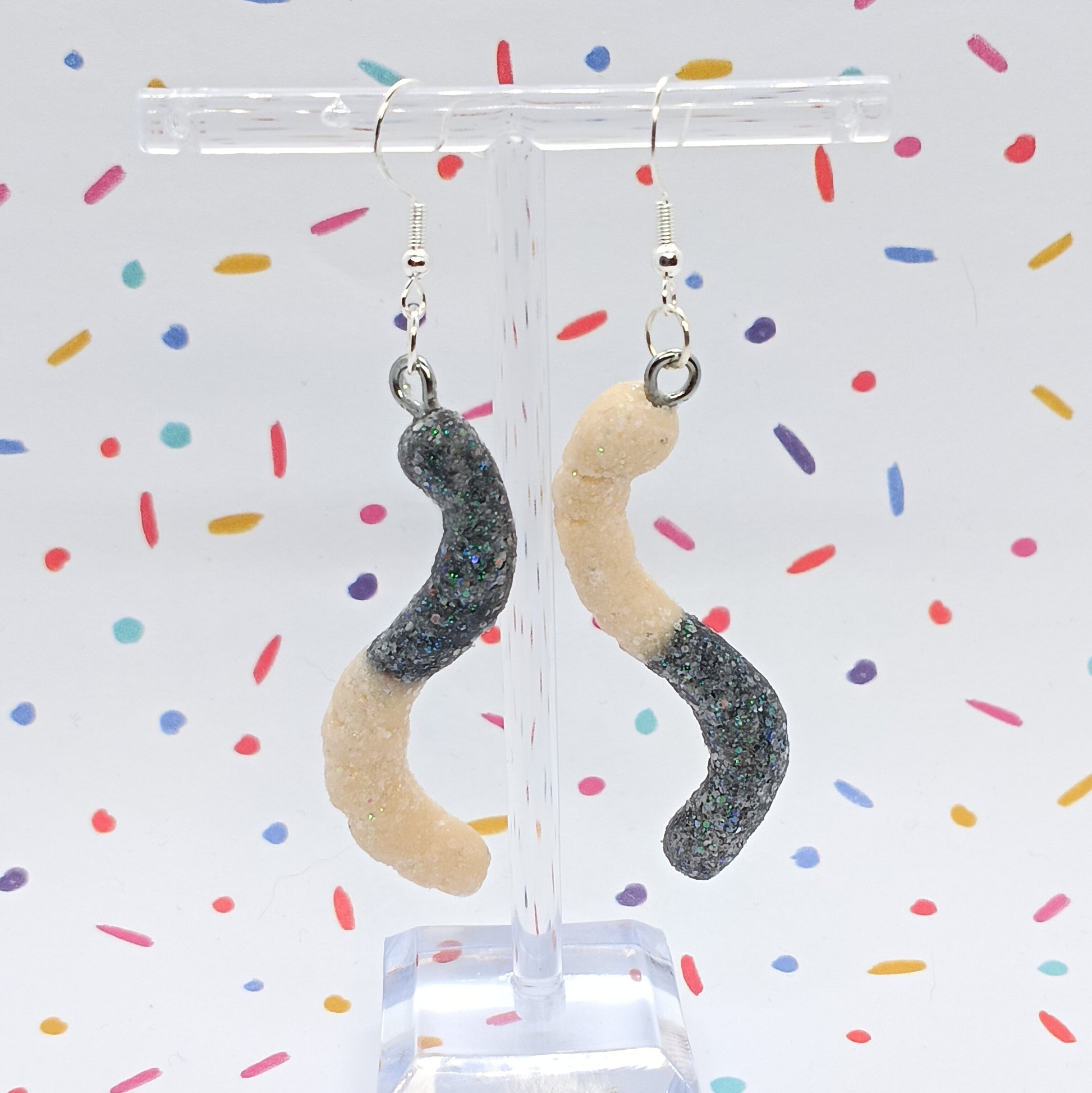 A pair of dangling earrings shaped like sour gummy worms, with glittery dark gray and beige sections, hanging from a clear acrylic display stand against a white background with colorful confetti.