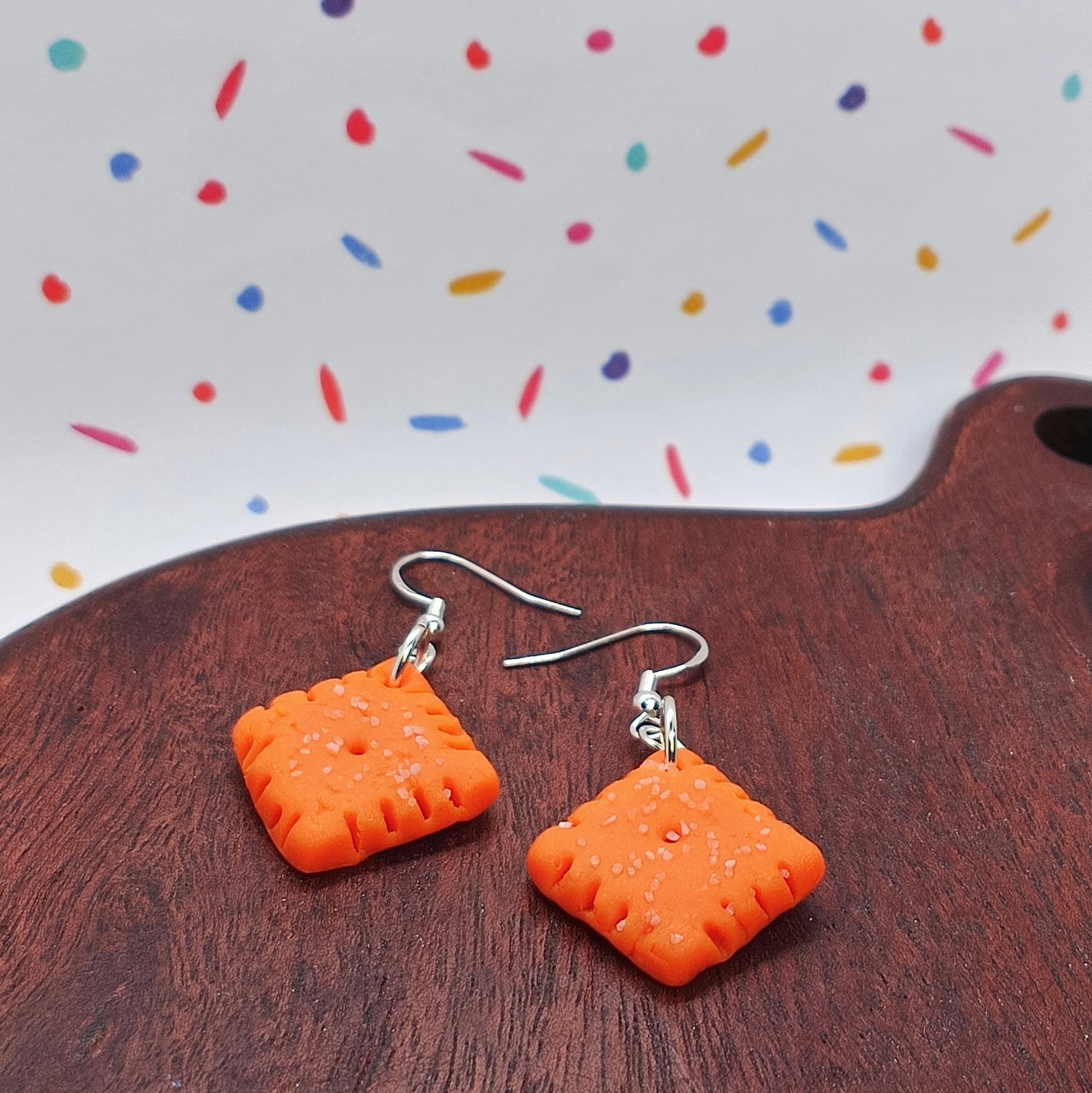 A pair of bright orange square dangle earrings with silver fishhook wires, displayed on a dark wooden surface with a white background featuring colorful confetti-like specks.