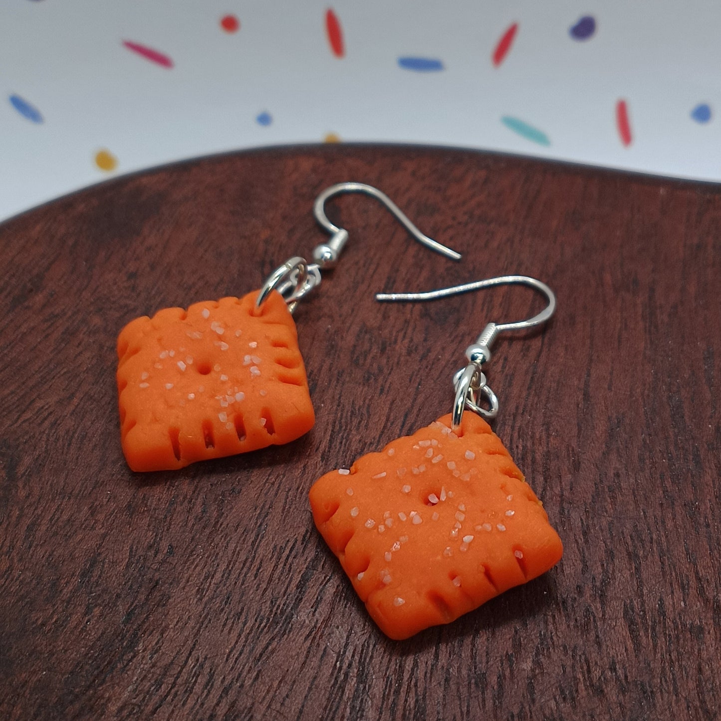 Orange polymer clay drop earrings shaped like square crackers with silver fishhook wires, displayed on a dark brown wooden surface.