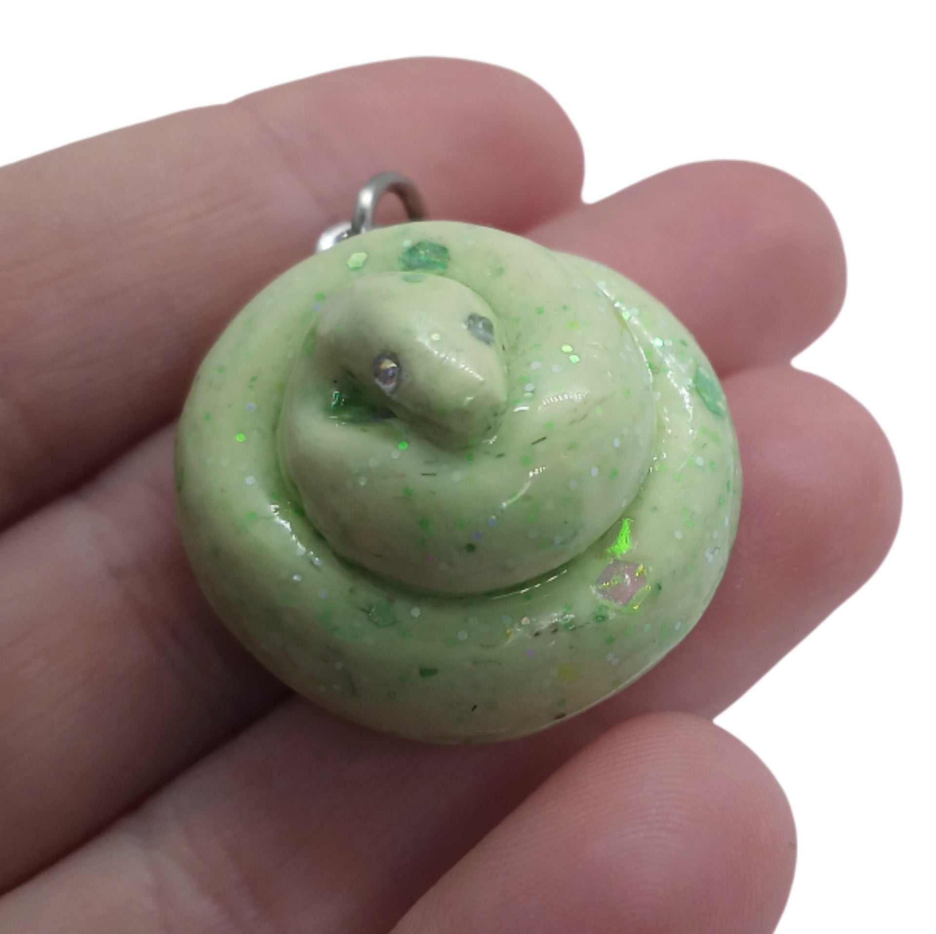 A small coiled snake charm or pendant in glossy light green with tiny glitter flecks and small silver eyes, resting on a person's fingers against a white background.