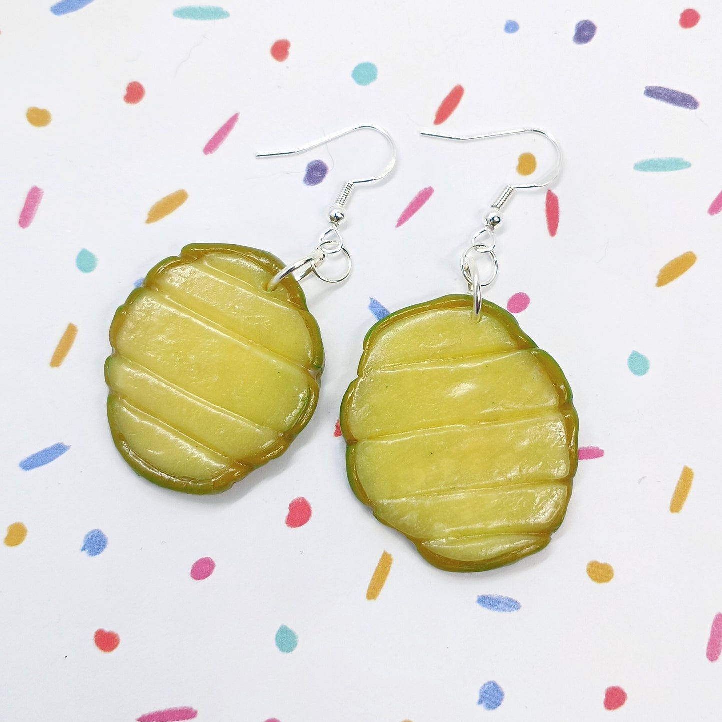 A pair of handmade earrings shaped like pickle slices, featuring a textured olive-green and yellow-green design with silver fishhook wires, displayed on a white background with colorful confetti dots.