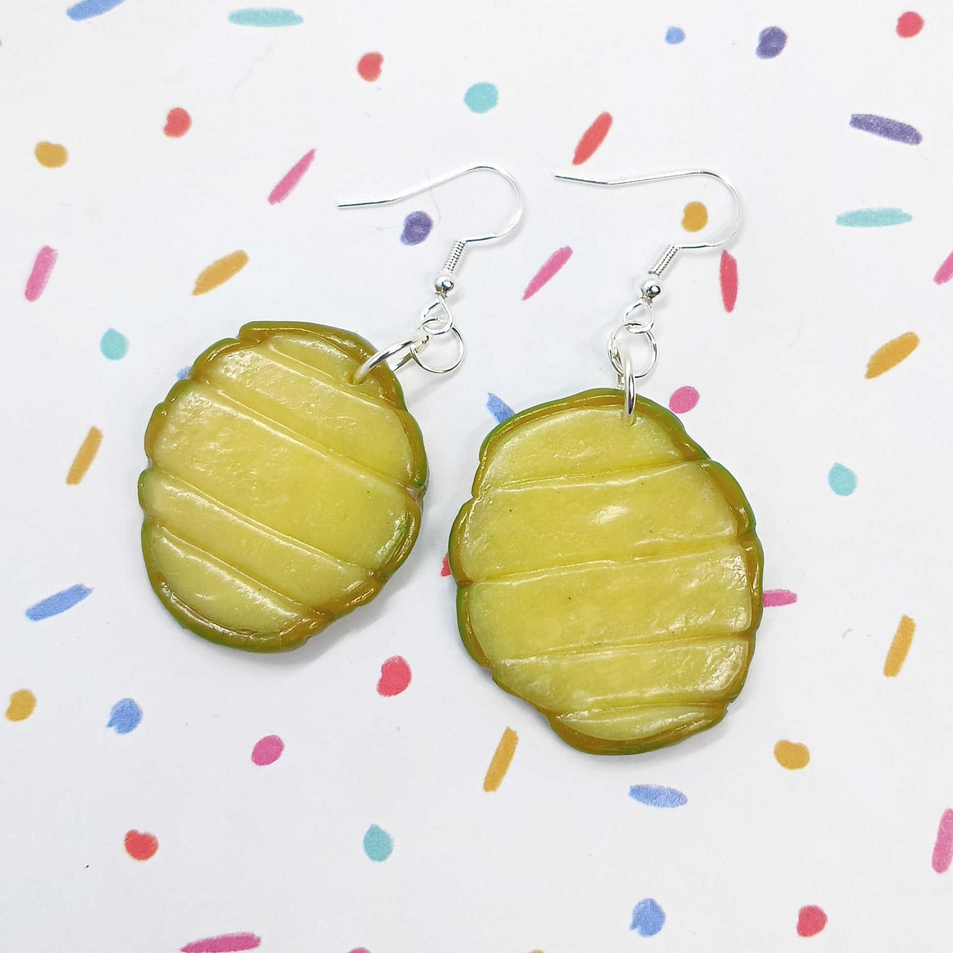 A pair of handmade earrings shaped like pickle slices, featuring a textured olive-green and yellow-green design with silver fishhook wires, displayed on a white background with colorful confetti dots.