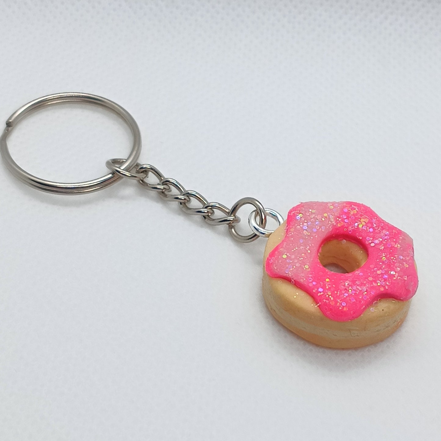 A novelty keychain featuring a small donut charm with pink glittery frosting and a silver metal split ring and chain, shown on a light textured background.