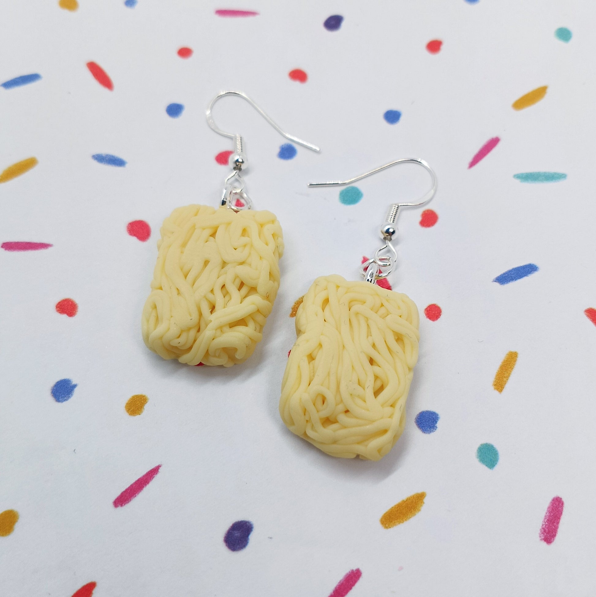 A pair of novelty dangle earrings shaped like yellow instant ramen noodles, hanging from silver fishhook wires on a white background with a colorful confetti pattern.