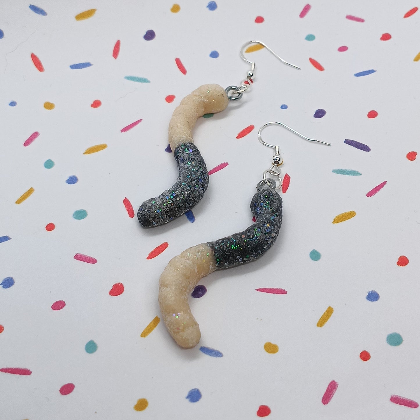 A pair of handmade worm-shaped dangle earrings with glittery black and beige bodies and silver fishhook wires, displayed on a white background with a colorful sprinkle pattern.
