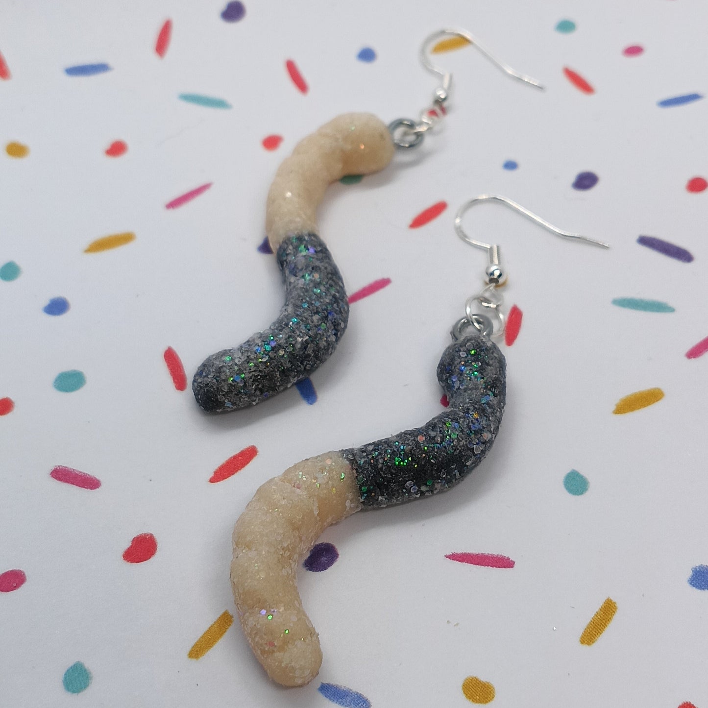 A pair of novelty worm-shaped dangle earrings with glittery black bodies and beige heads, hanging from silver fishhook wires on a white background with colorful confetti dots.