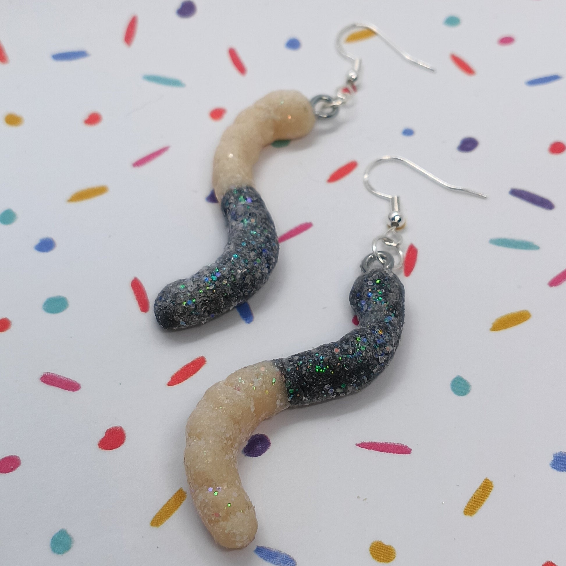 A pair of novelty worm-shaped dangle earrings with glittery black bodies and beige heads, hanging from silver fishhook wires on a white background with colorful confetti dots.