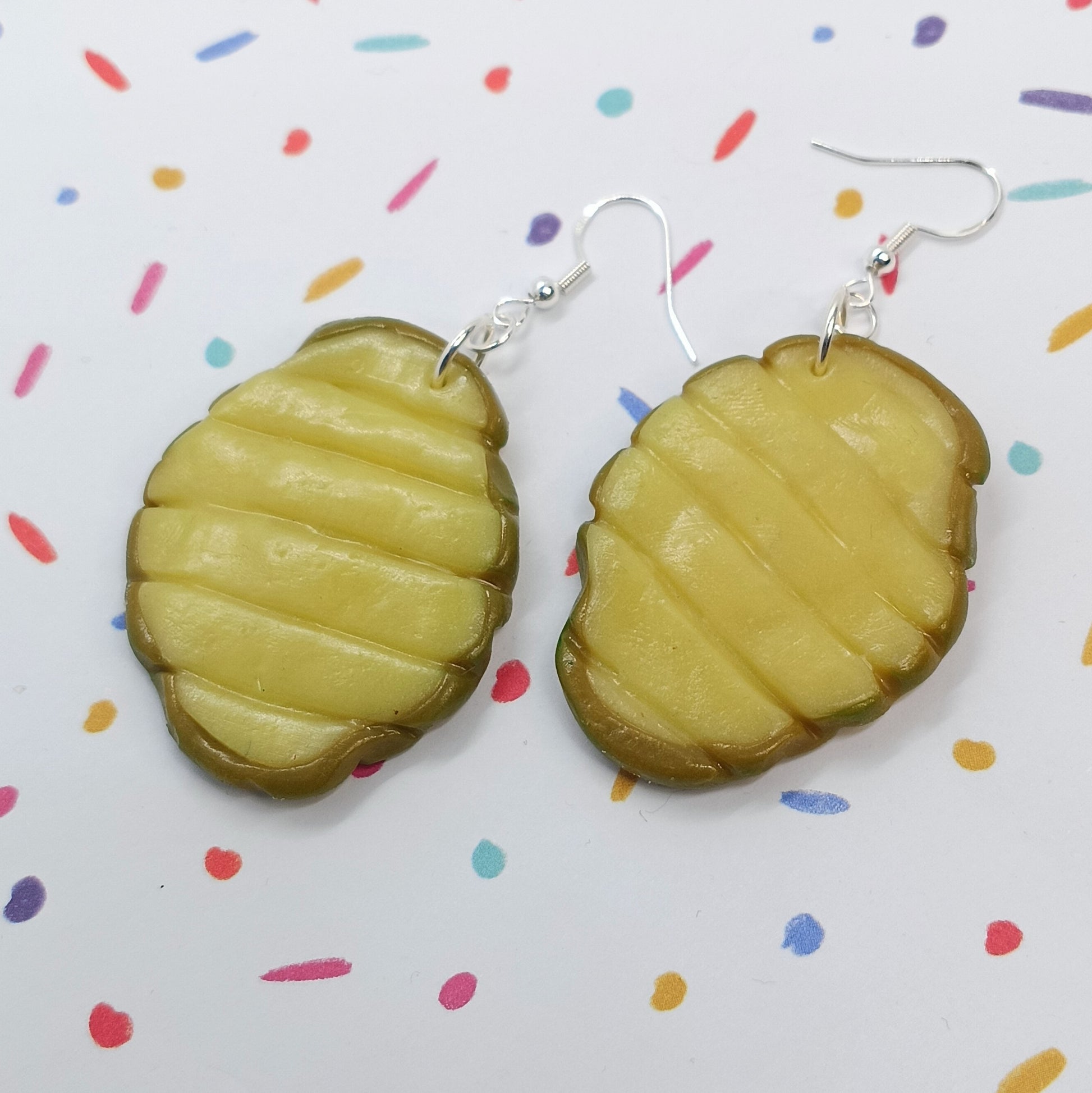 A pair of handmade polymer clay earrings shaped like pickle slices, featuring a yellow-green center and a dark green outer rind, suspended from silver fishhook wires on a white background with colorful confetti dots.