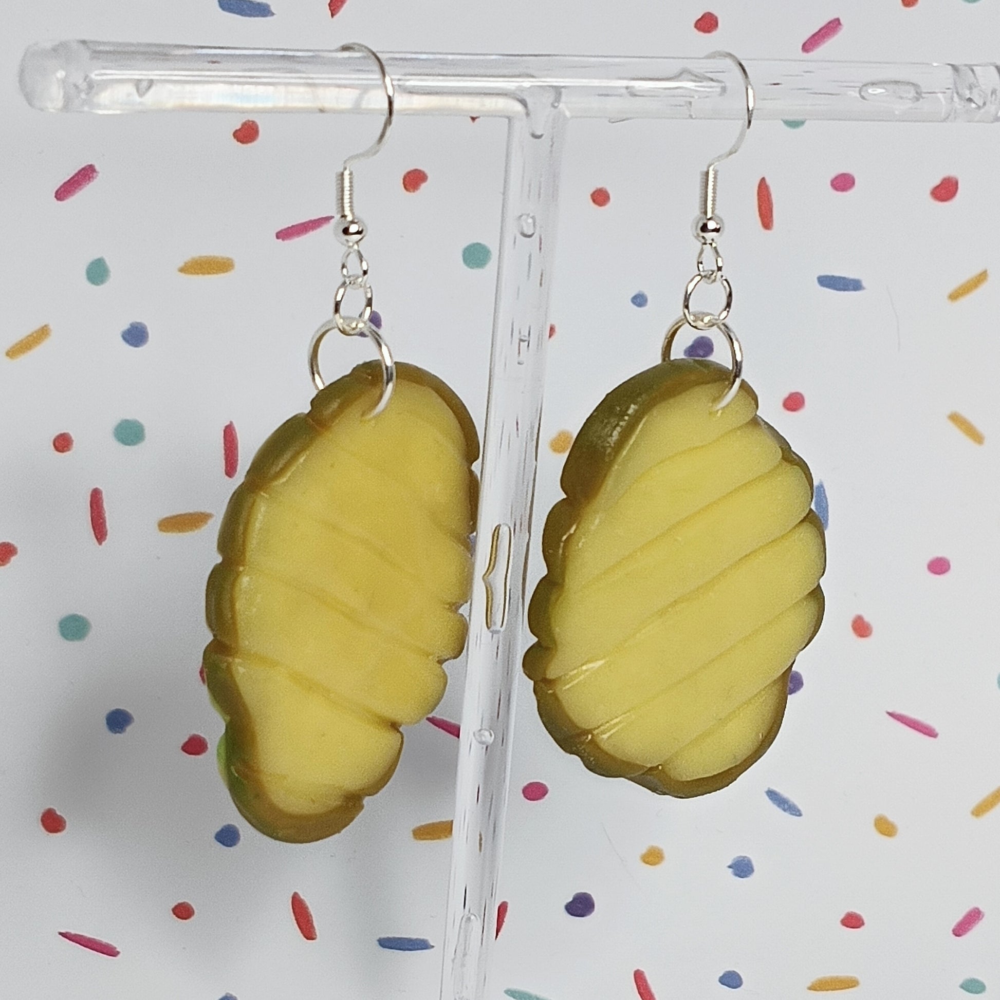 A pair of novelty earrings shaped like sliced pickles, hanging from silver fishhook wires on a clear acrylic display stand against a white background with colorful confetti.