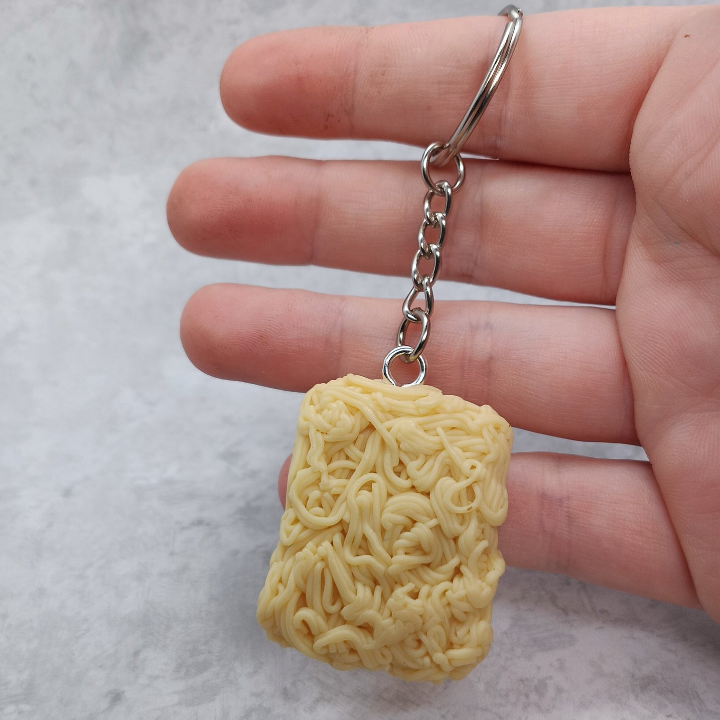 A miniature yellow ramen noodle keychain held in a person's hand against a light gray textured background.
