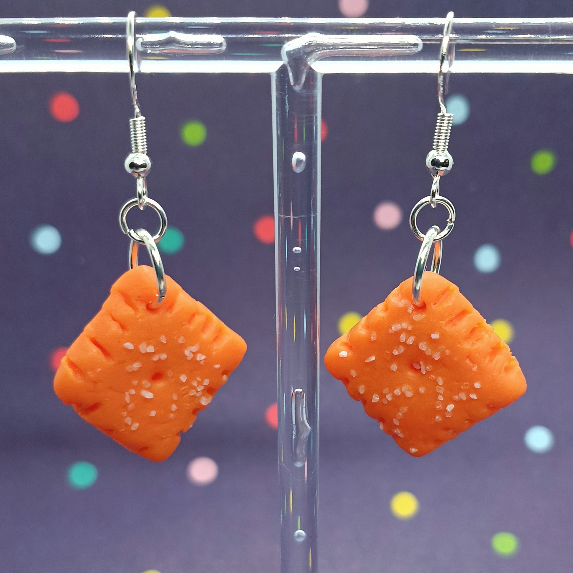 A pair of orange square dangle earrings with white salt-like speckles, hanging from silver fishhook wires on a clear acrylic display stand against a dark purple background with colorful confetti.