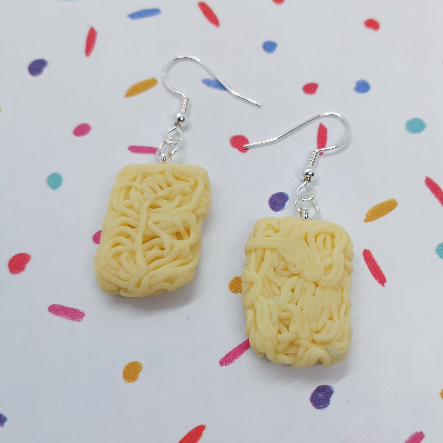 A pair of novelty dangle earrings shaped like mini instant ramen noodle packs, suspended from silver fishhook wires, displayed on a white background with a colorful confetti pattern.