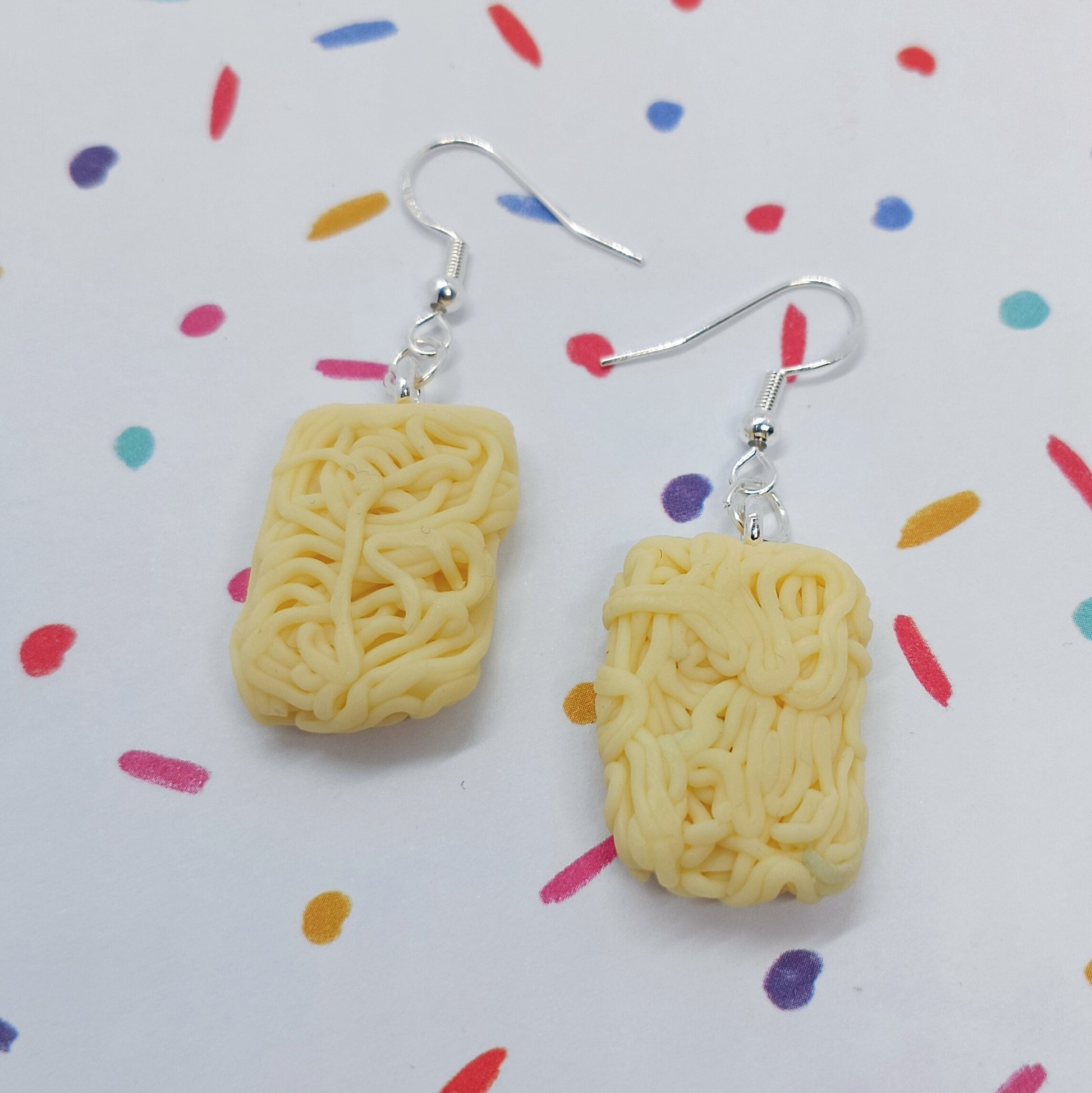 A pair of novelty dangle earrings shaped like mini instant ramen noodle packs, suspended from silver fishhook wires, displayed on a white background with a colorful confetti pattern.
