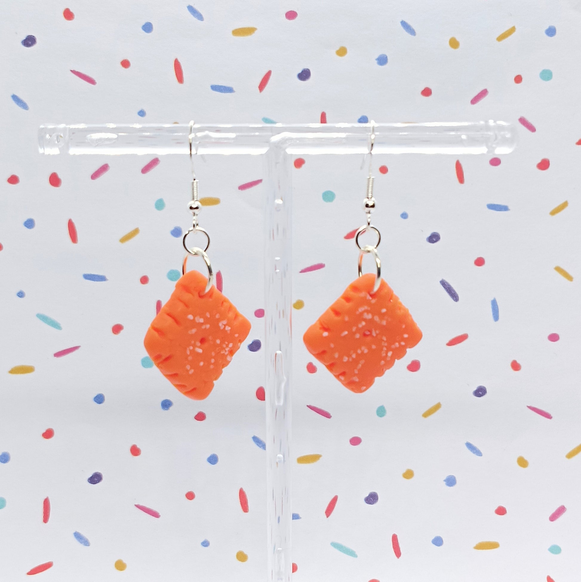 A pair of orange square-shaped dangling earrings with small white speckles, hanging from silver hooks on a clear T-stand against a white background with colorful confetti.