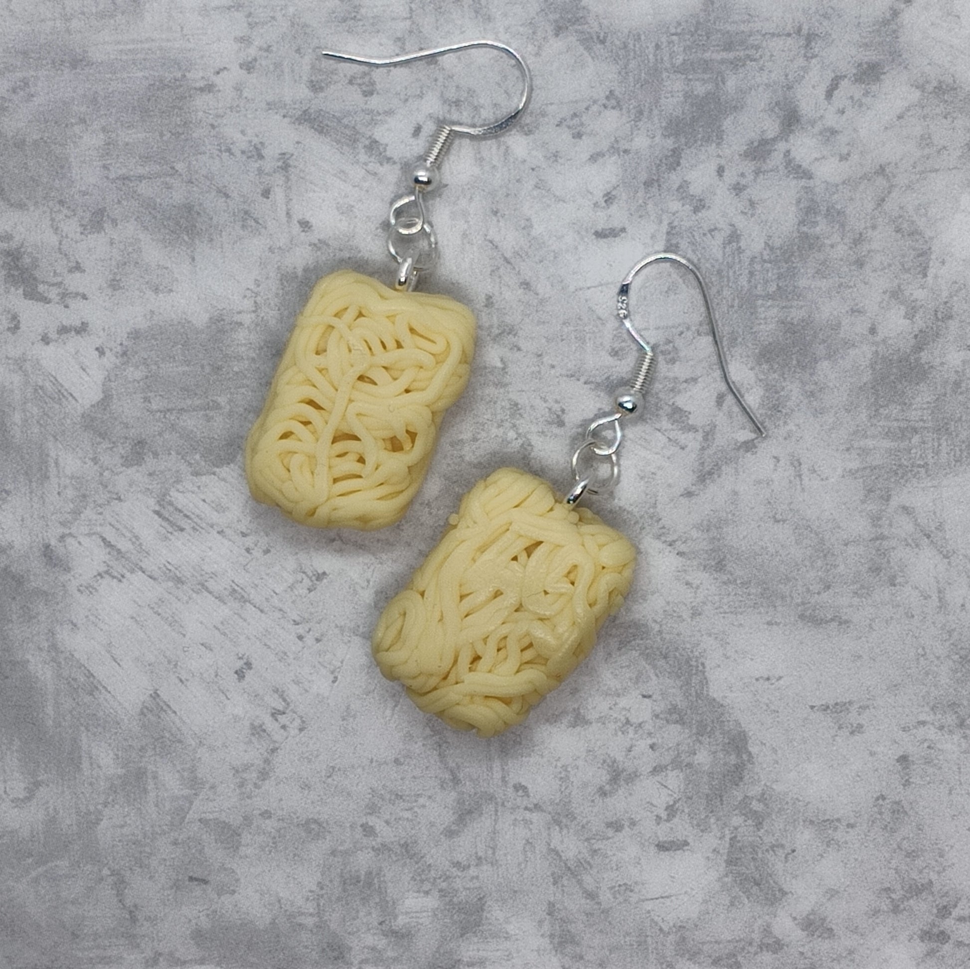 A pair of handmade dangle earrings featuring textured, sculpted pale yellow pasta shapes hanging from silver fishhook wires, displayed on a textured gray background.