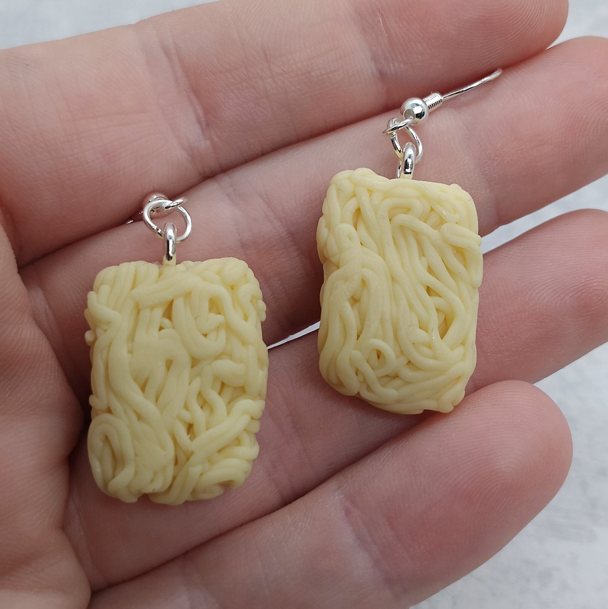 A pair of novelty dangle earrings shaped like a block of instant ramen noodles, held in a person's hand. The earrings are pale yellow with silver fishhook wires.