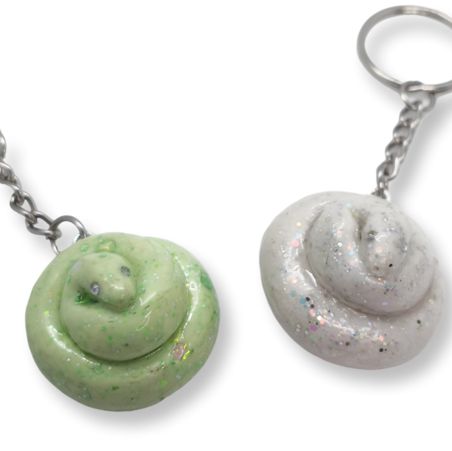 Two novelty keychains shaped like coiled poop, shown on a white background. One keychain is light green with green glitter, and the other is light gray with silver glitter.
