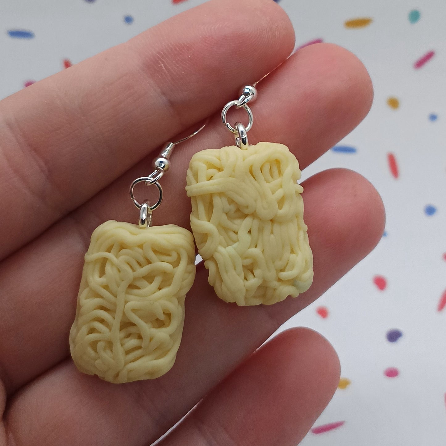 A pair of novelty pasta-shaped drop earrings with silver fishhook wires, held in a person's hand against a white background with colorful confetti-like specks.