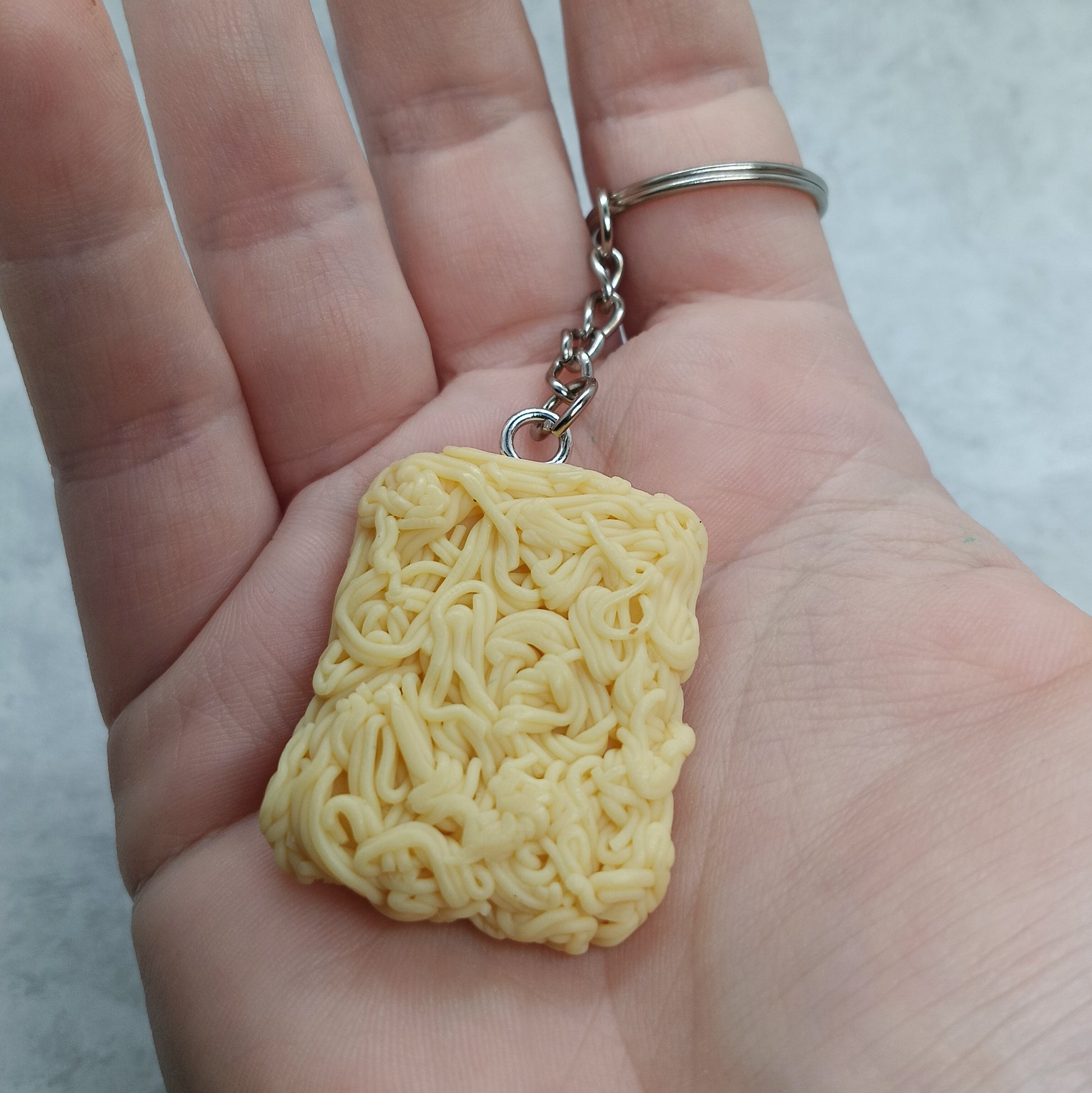 A miniature yellow ramen noodle keychain held in a person's hand. The keychain features a highly detailed, textured yellow noodle charm attached to a silver metal split ring and short chain.
