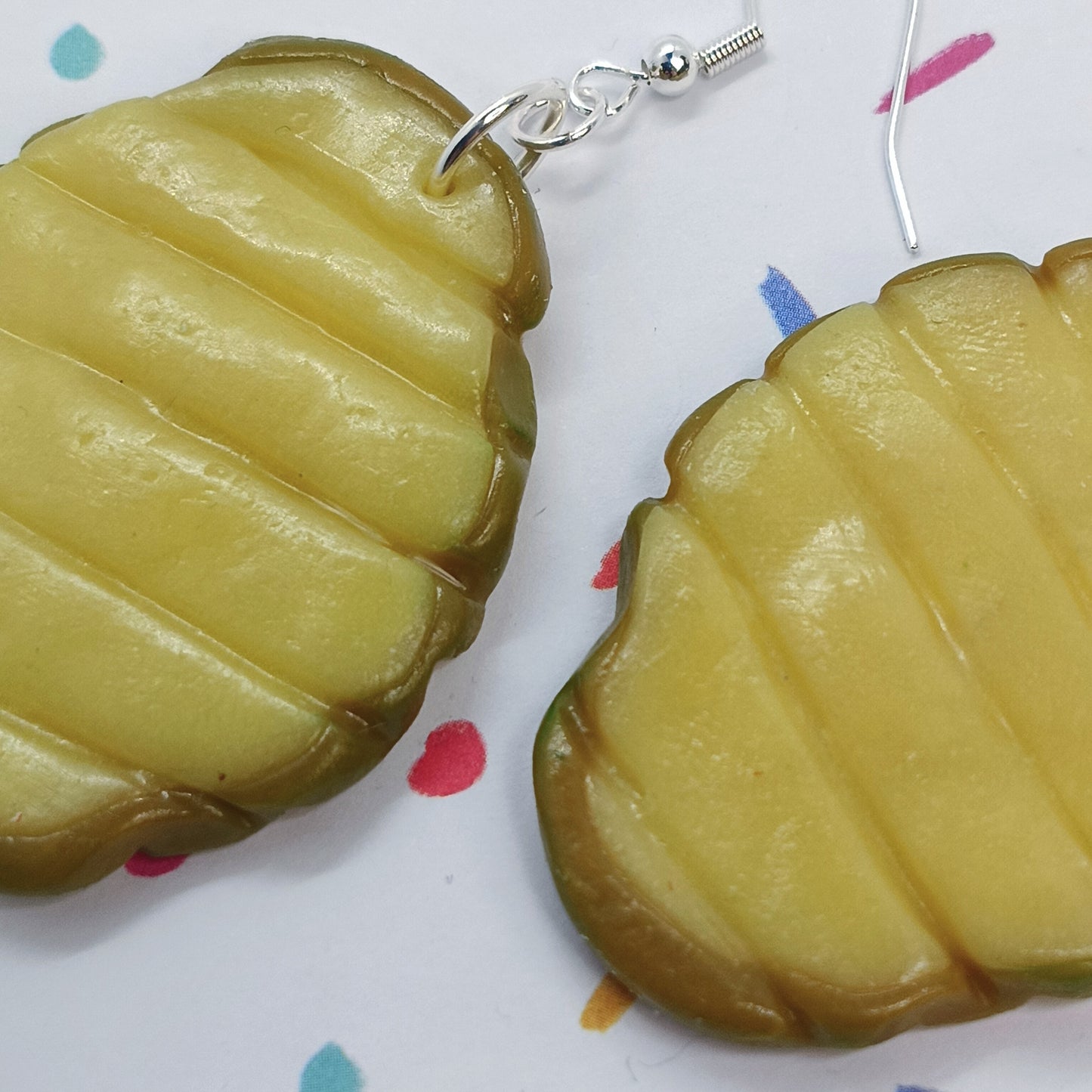 Pickle-shaped dangle earrings with a glossy yellow-green surface and silver fishhook wires, shown on a white background with small colorful confetti dots.