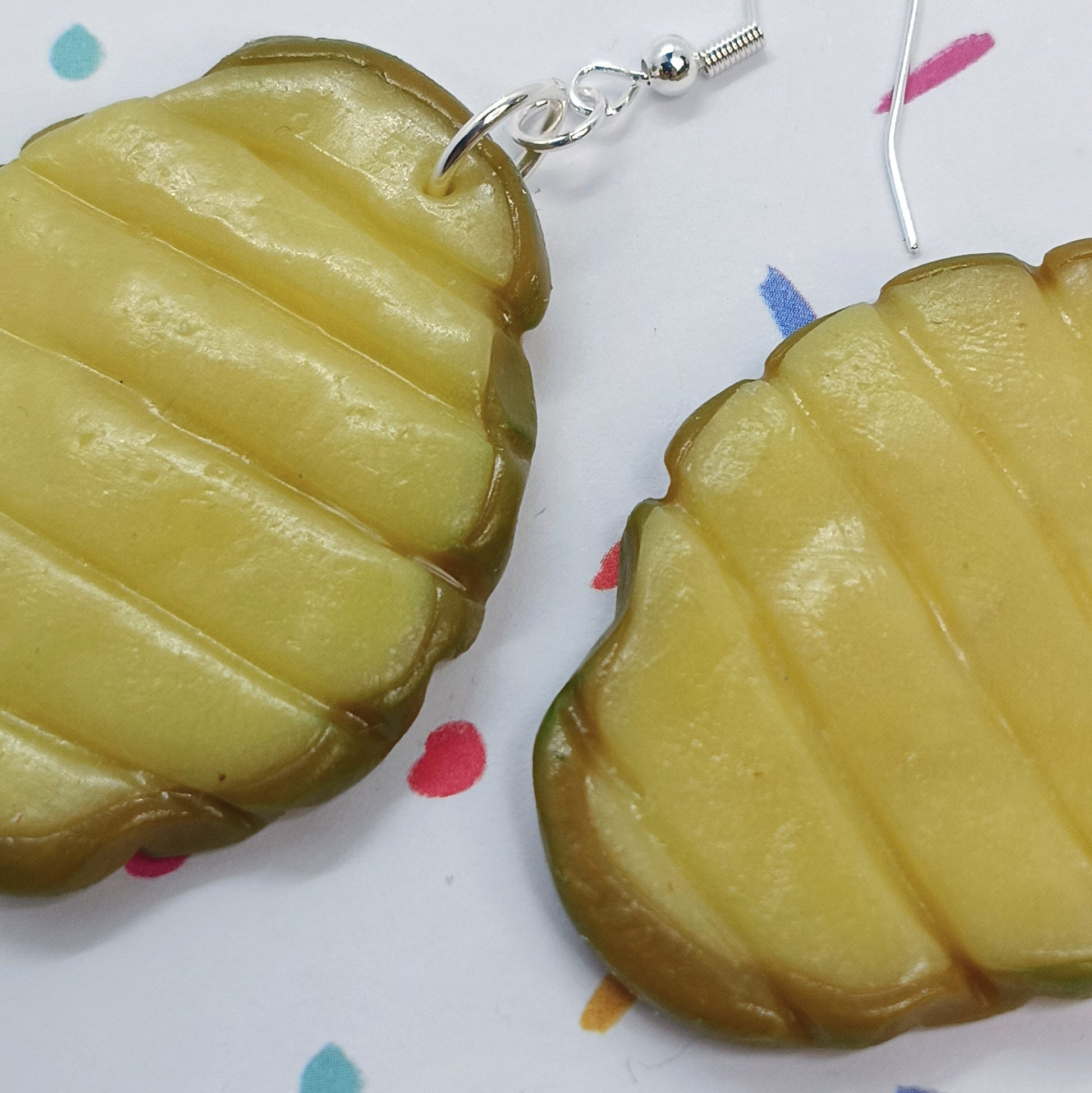 Pickle-shaped dangle earrings with a glossy yellow-green surface and silver fishhook wires, shown on a white background with small colorful confetti dots.