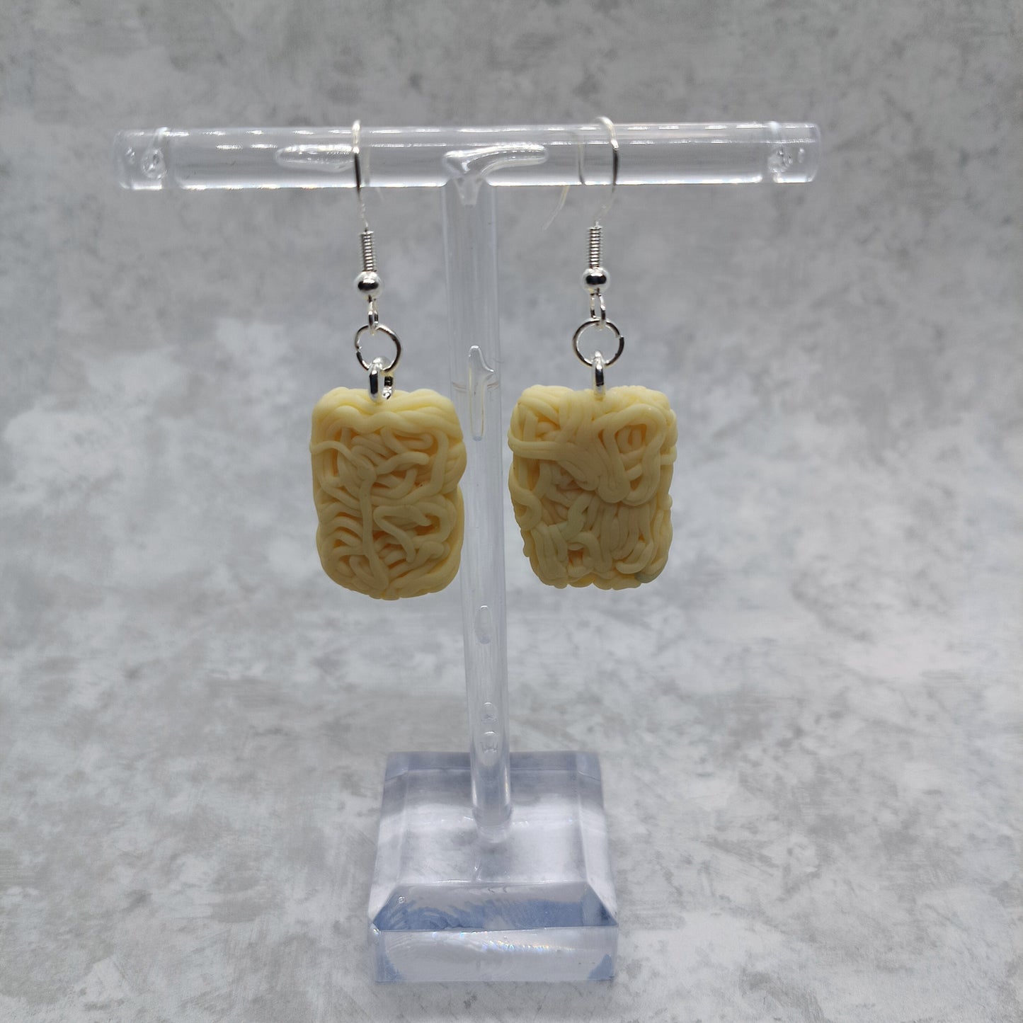 A pair of ramen pale yellow rectangular drop earrings with silver fishhook wires, displayed on a clear acrylic stand against a textured gray background.