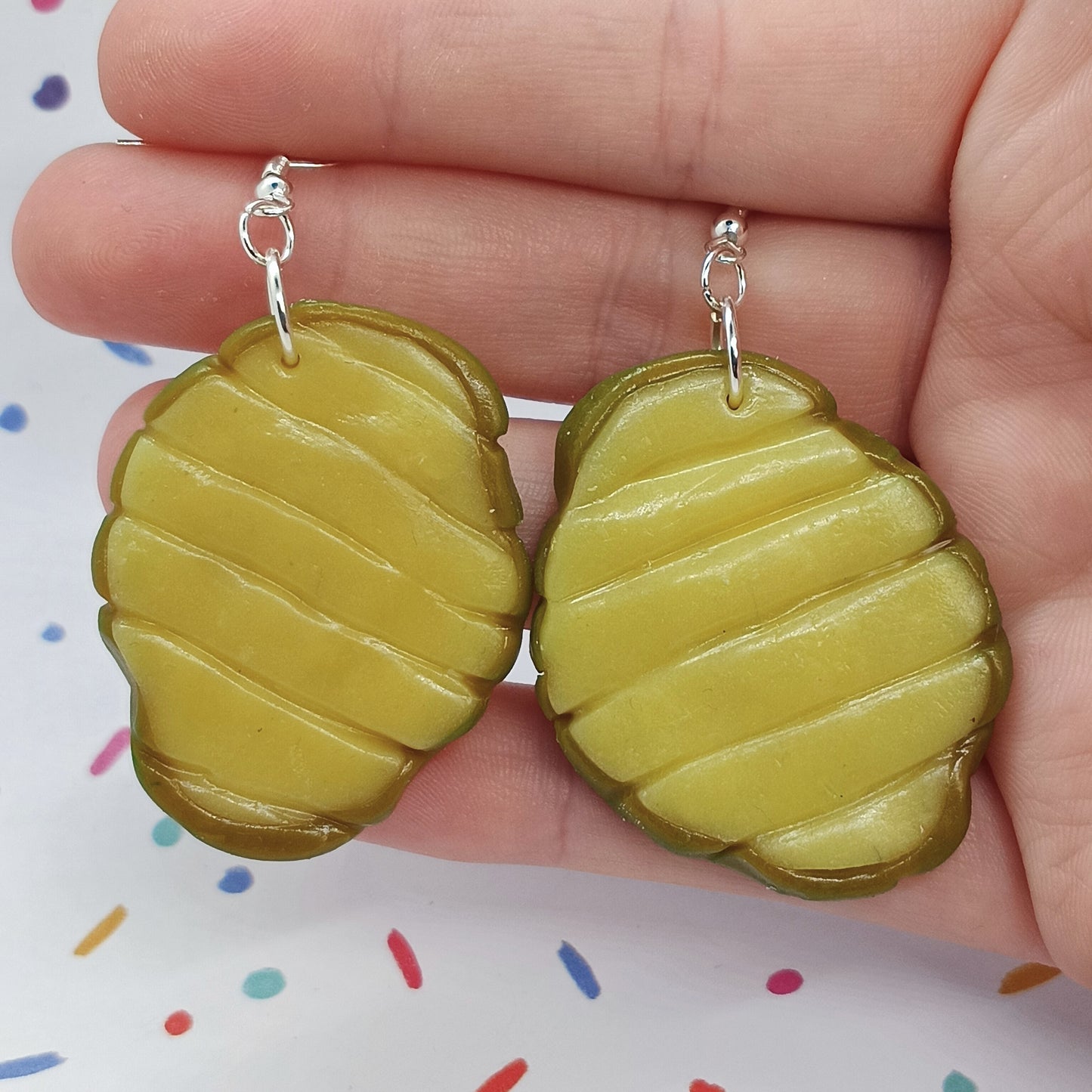 A pair of pickle-shaped earrings with a glossy yellow-green surface and silver fishhook wires, held in a person's hand against a white background with colorful confetti.