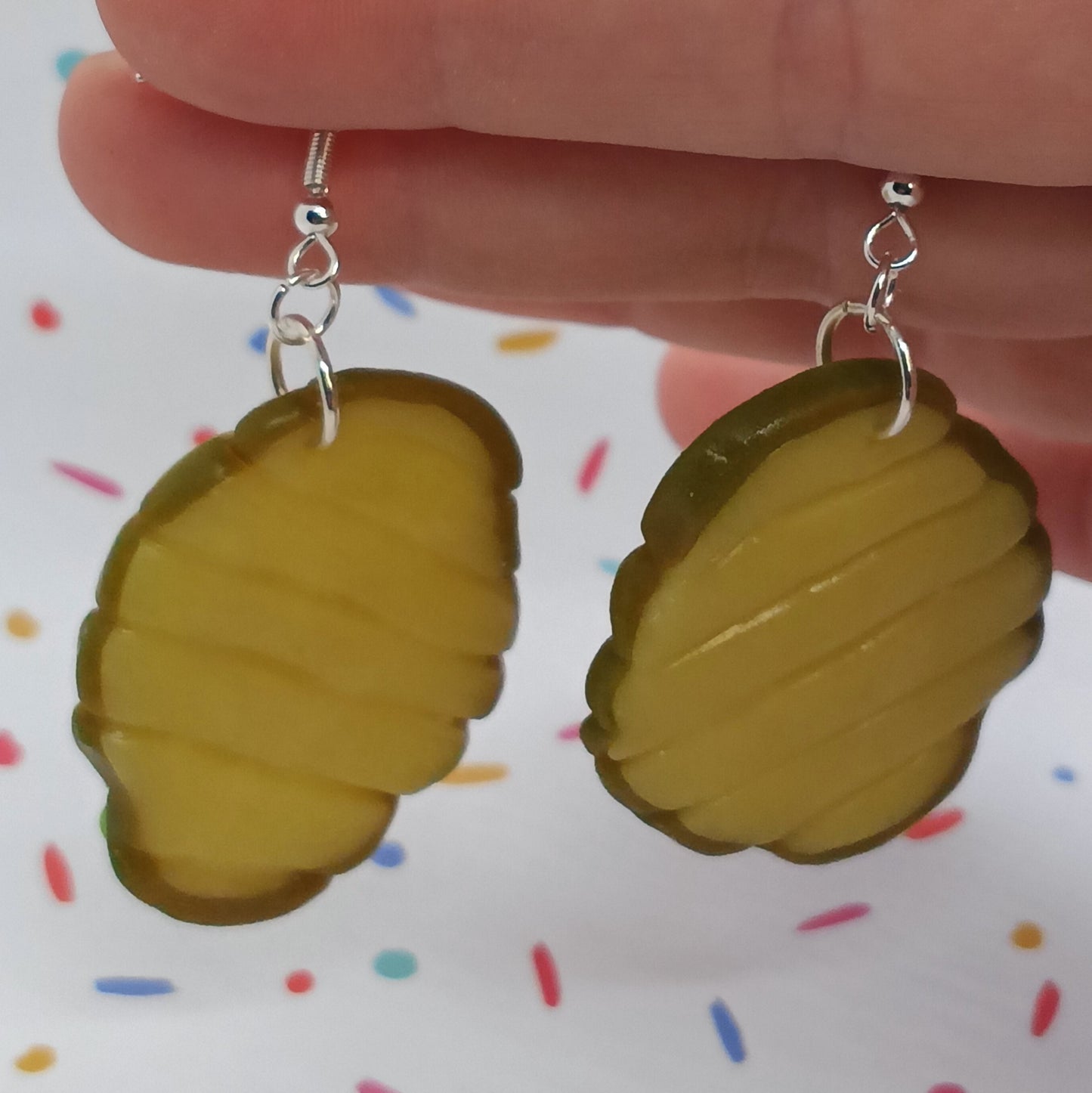 A pair of pickle-shaped drop earrings with silver fishhook wires, held by a hand against a white background with colorful confetti.