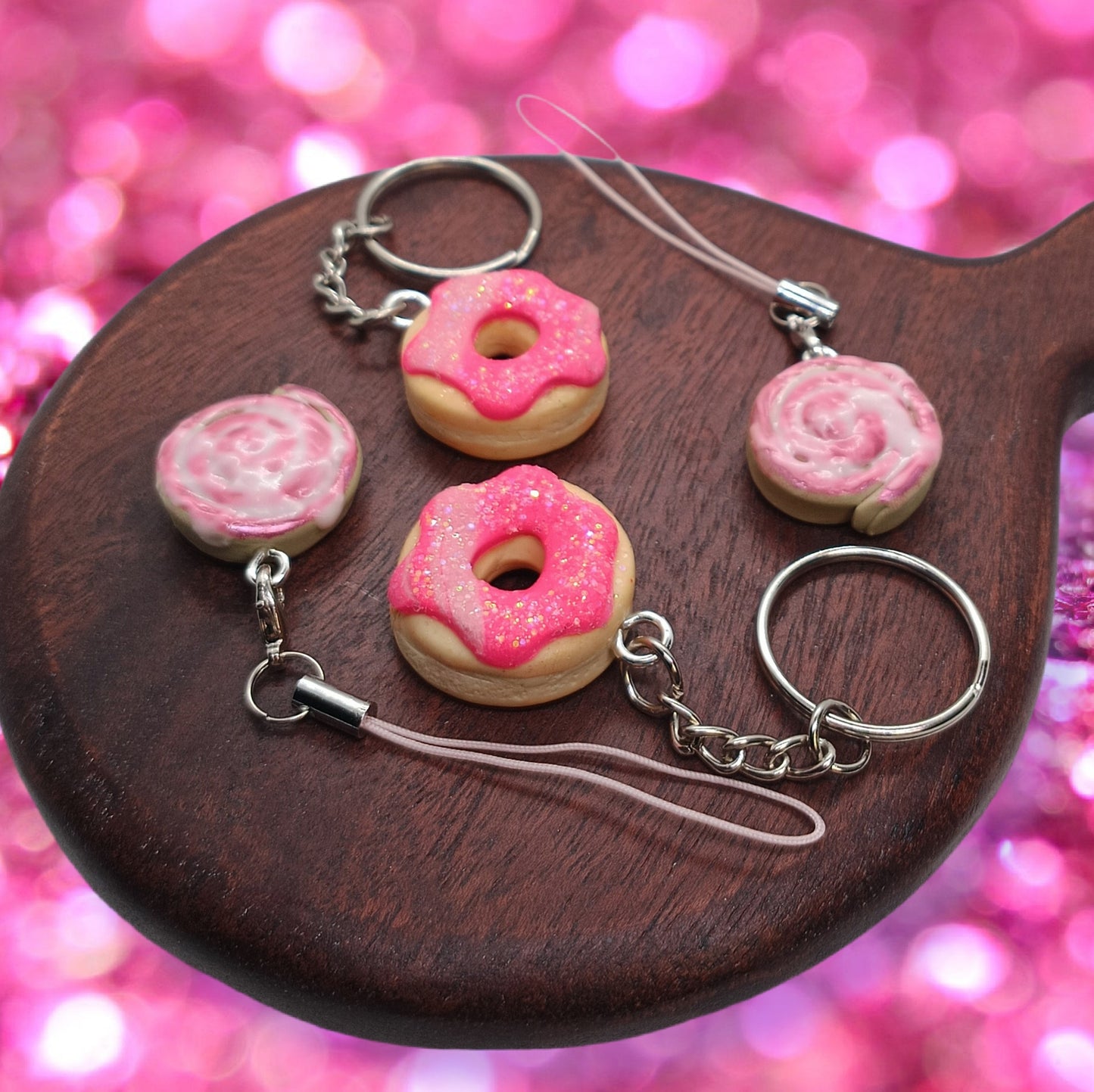 Four small keychain charms displayed on a dark wooden spoon against a pink glittery bokeh background. The set includes two pink-frosted donut keychains with silver metal rings and two pink-and-white swirl cinnamon roll charms.