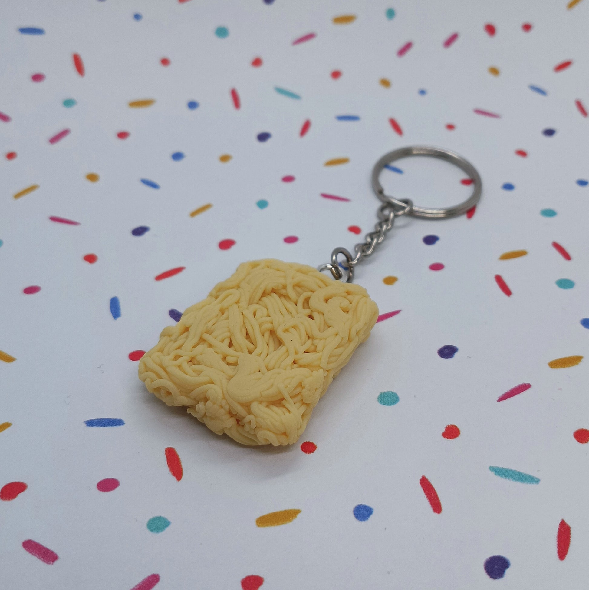 A novelty keychain shaped like a block of ramen noodles, attached to a silver metal split ring, set against a white background with colorful confetti sprinkles.